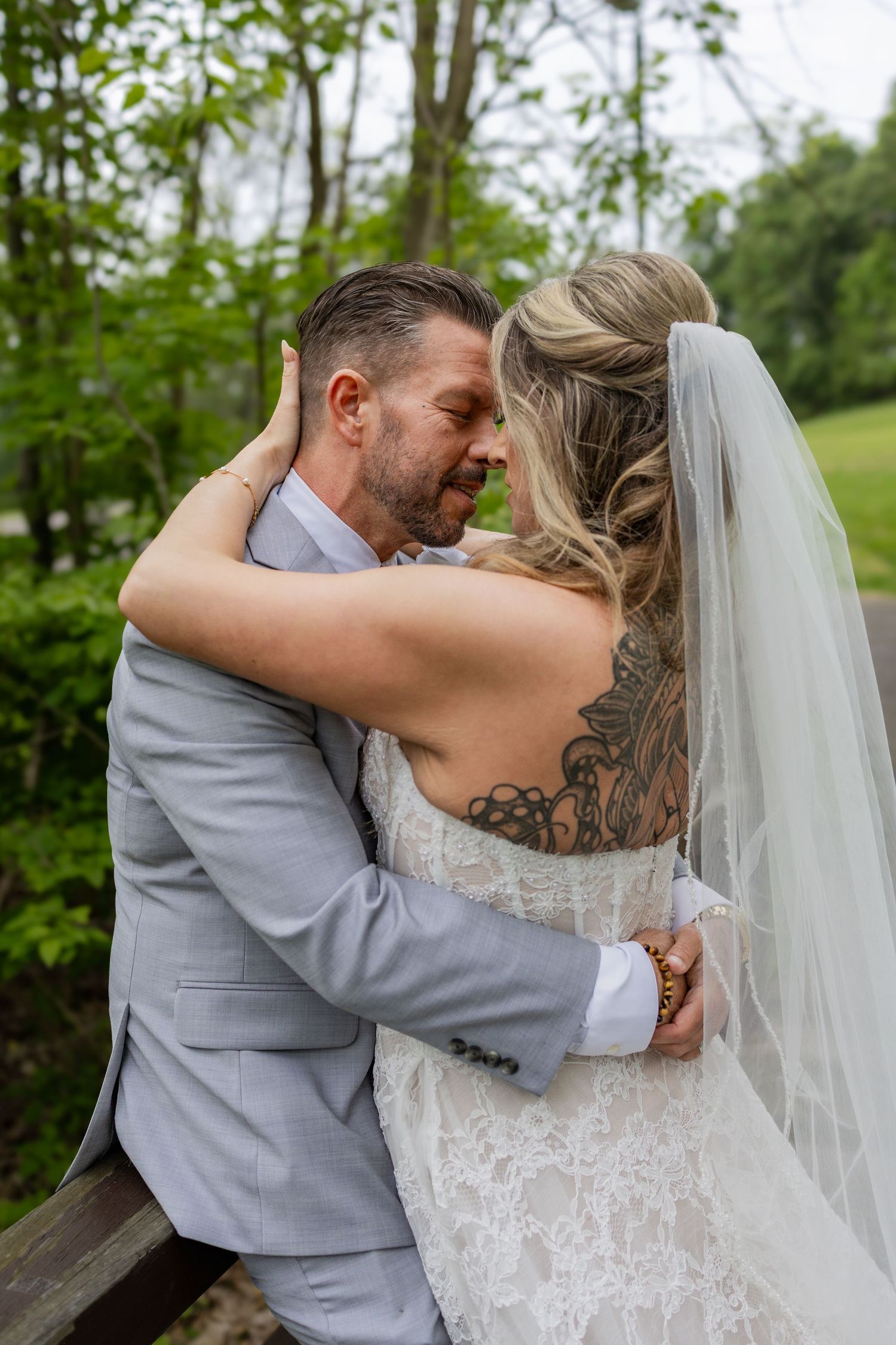 A bride with a back tattoo and veil embraces her groom in a gray suit outdoors at their wedding.