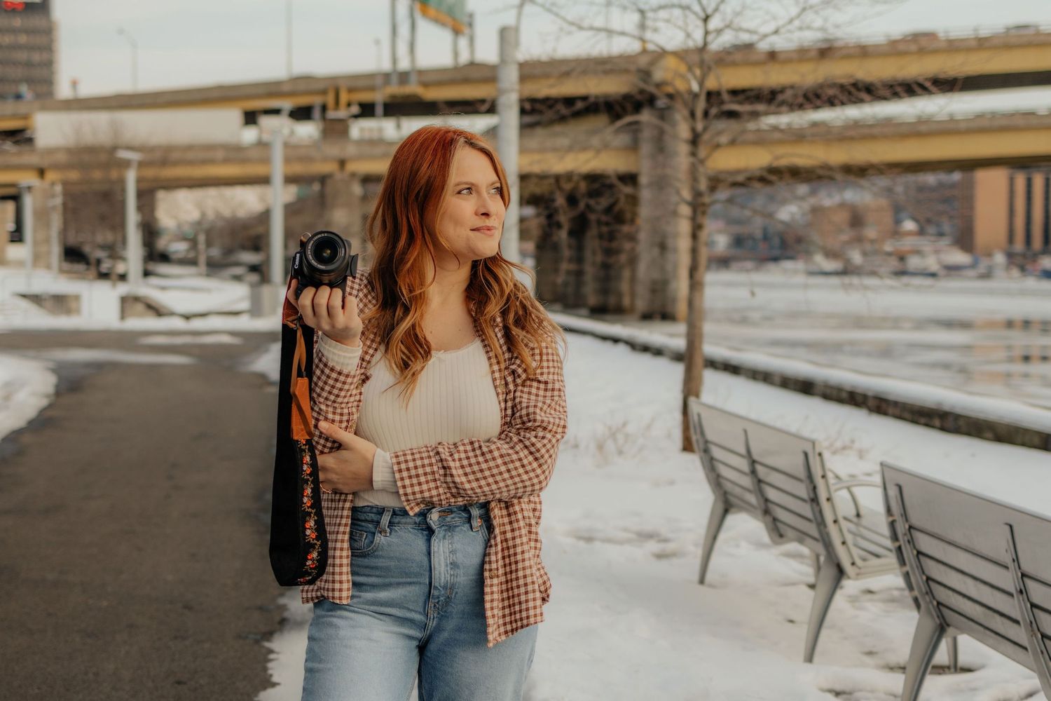 Red-haired woman holding a camera outdoors in winter, wearing a plaid jacket and jeans near a bridge.