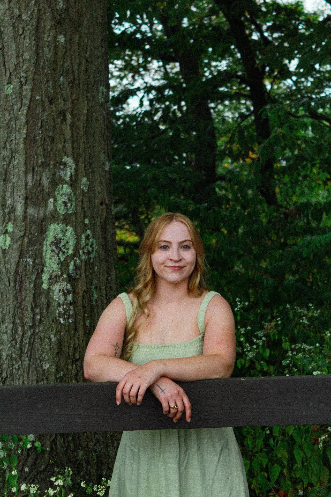 Young woman in green dress leaning on wooden fence beside large tree in lush garden setting.