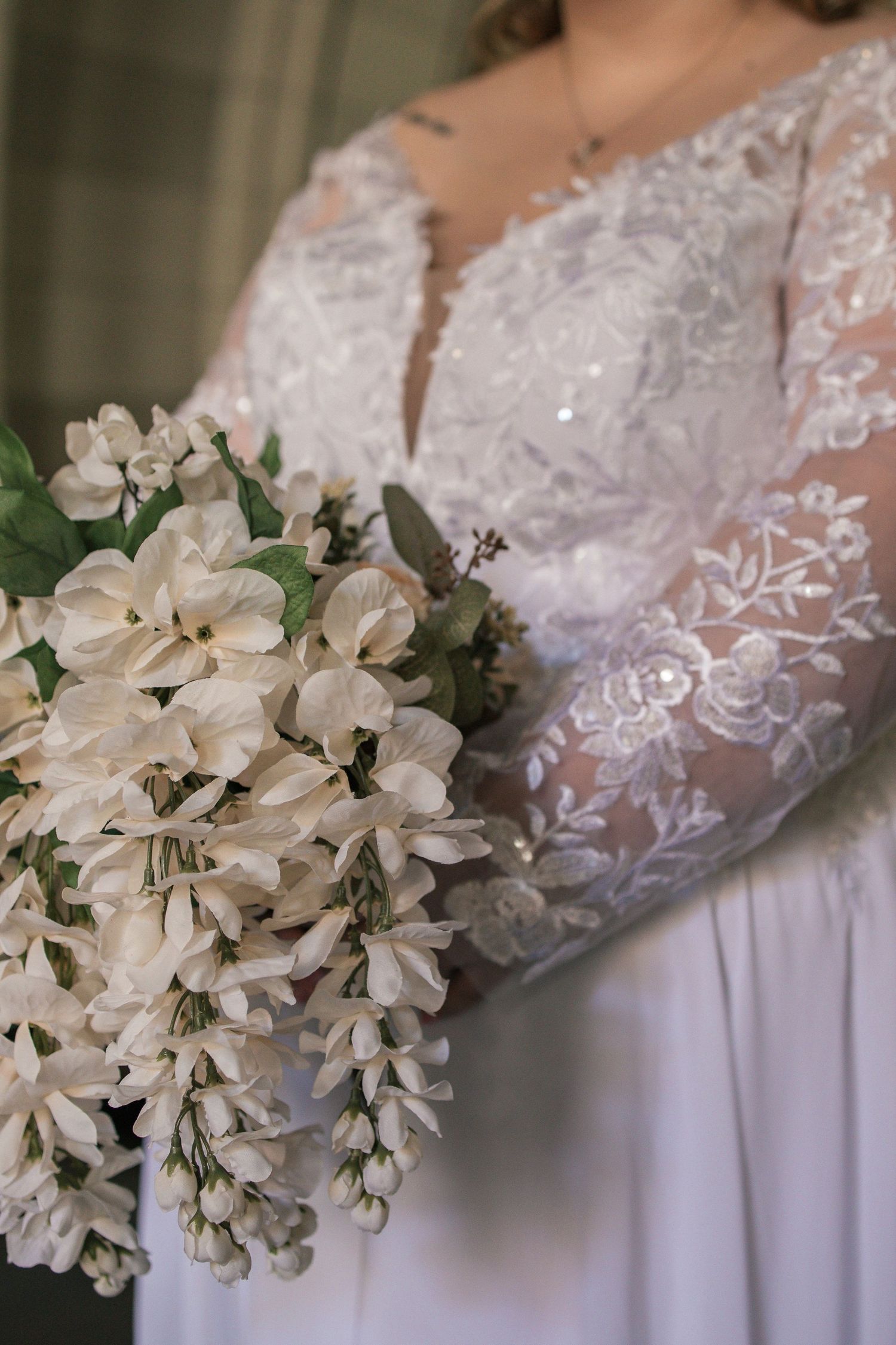 Bride in lace long-sleeve wedding gown holding elegant white floral bouquet with cascading flowers and greenery.