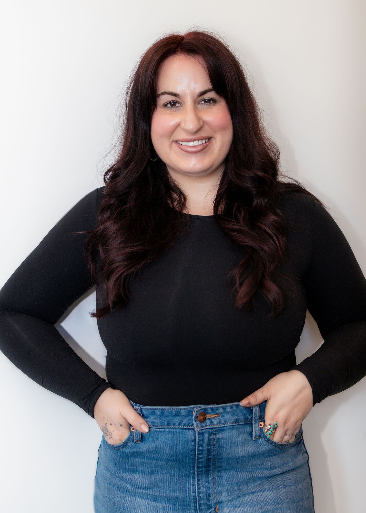 Woman with dark wavy hair smiling, wearing black long-sleeve top and denim skirt, posing with hands on hips.
