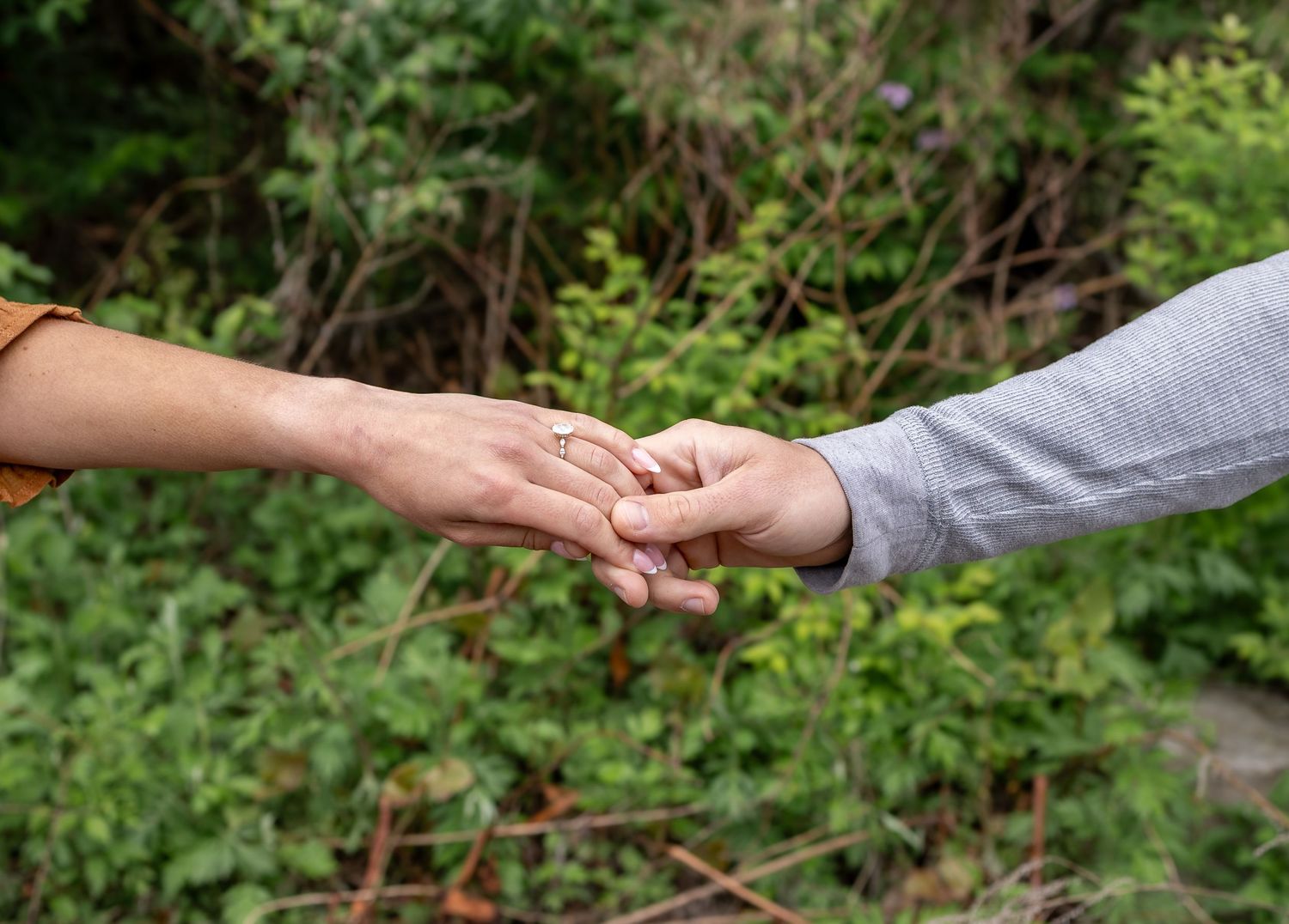 Two people gently holding hands outdoors with lush green foliage in the background.