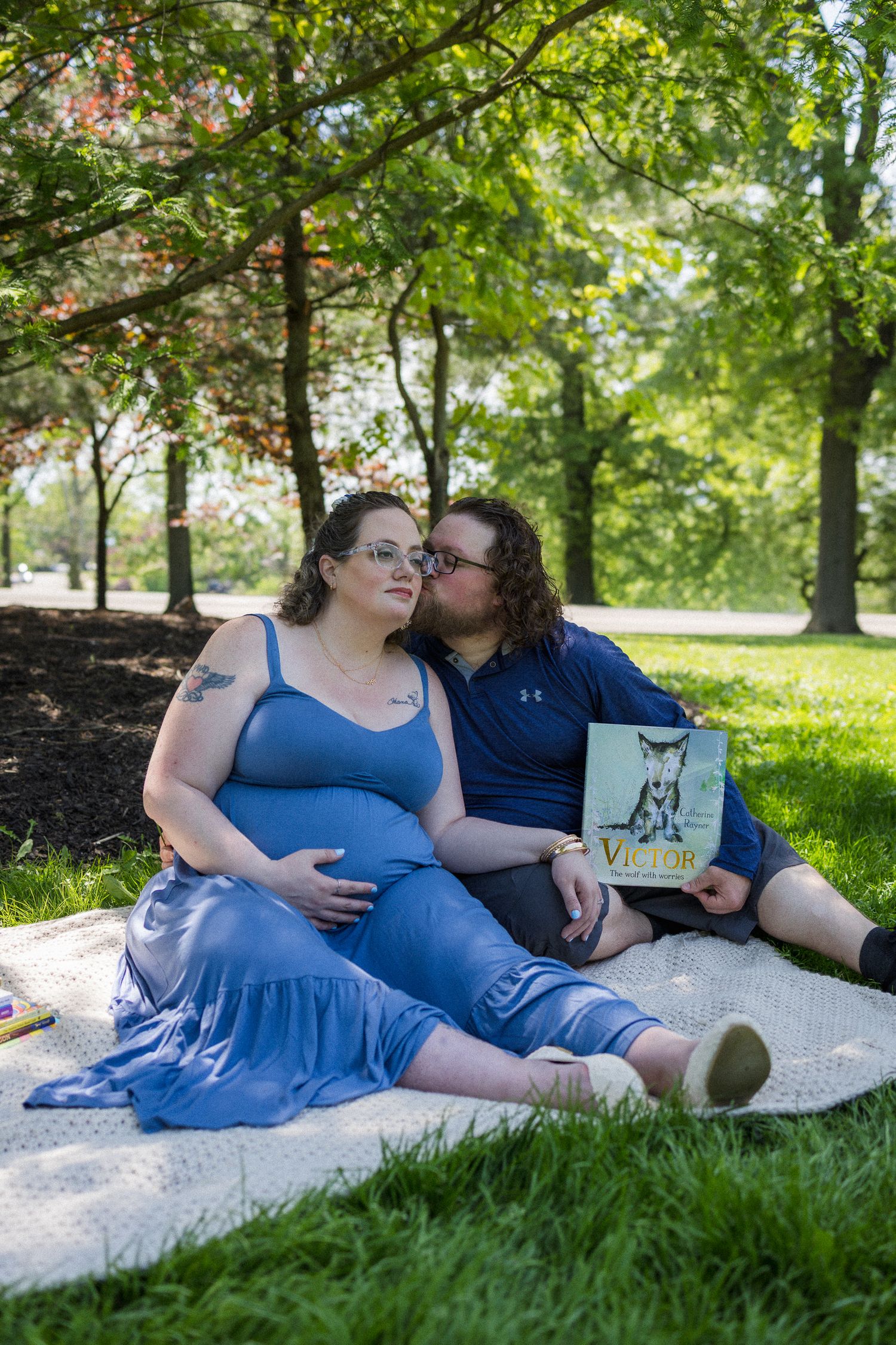 Pregnant couple sitting on blanket in park, man kissing woman's cheek while holding a children's book outdoors.