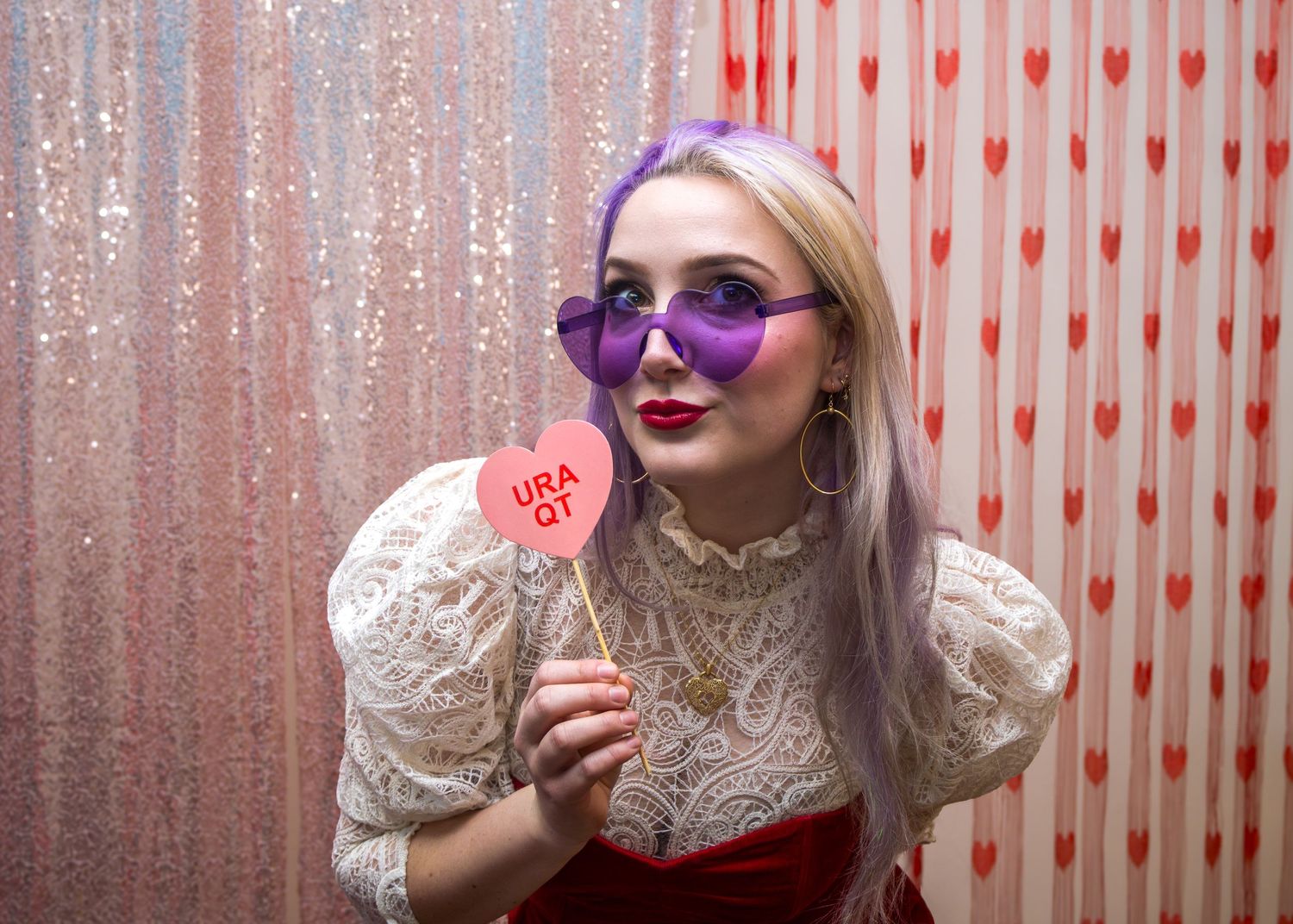 Woman with purple hair and heart-shaped sunglasses holds a Valentine candy heart prop in front of a festive backdrop.