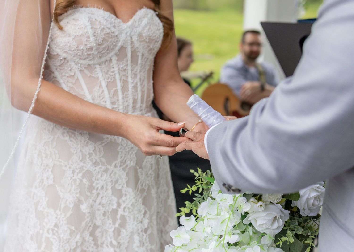 Bride in lace wedding gown exchanging rings with groom during outdoor wedding ceremony with guitarist in background.