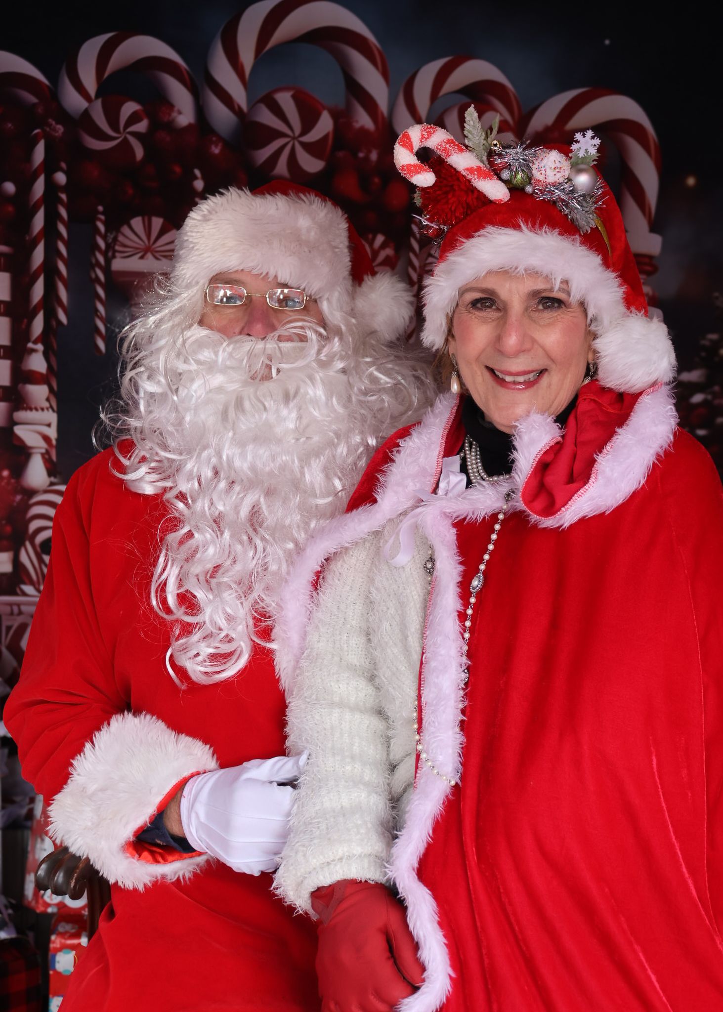 Santa Claus and Mrs. Claus pose together in festive red suits against a candy cane Christmas backdrop.