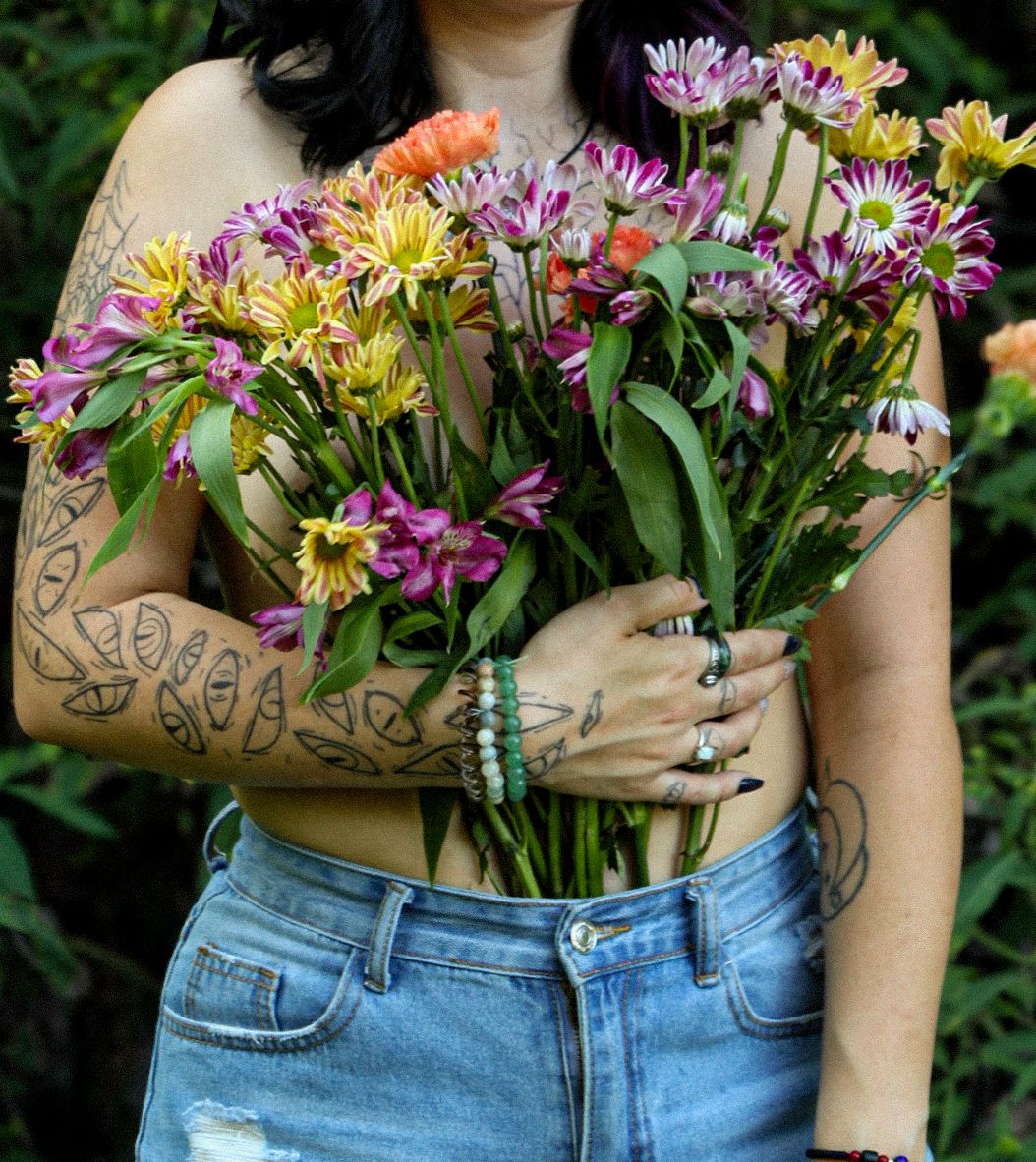 Woman with tattoos holding a vibrant bouquet of colorful daisies and wildflowers, wearing denim shorts and stacked bracelets.
