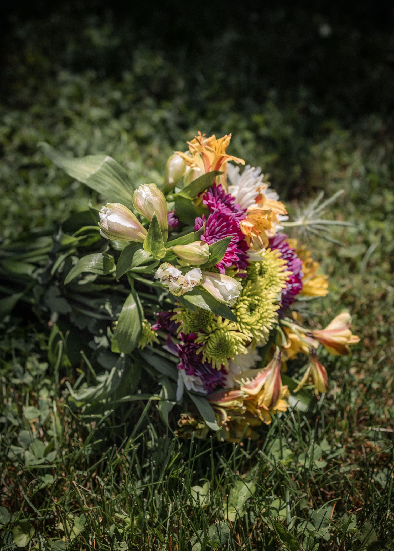 Colorful flower bouquet with pink, yellow, and orange blooms resting on green grass in natural sunlight.