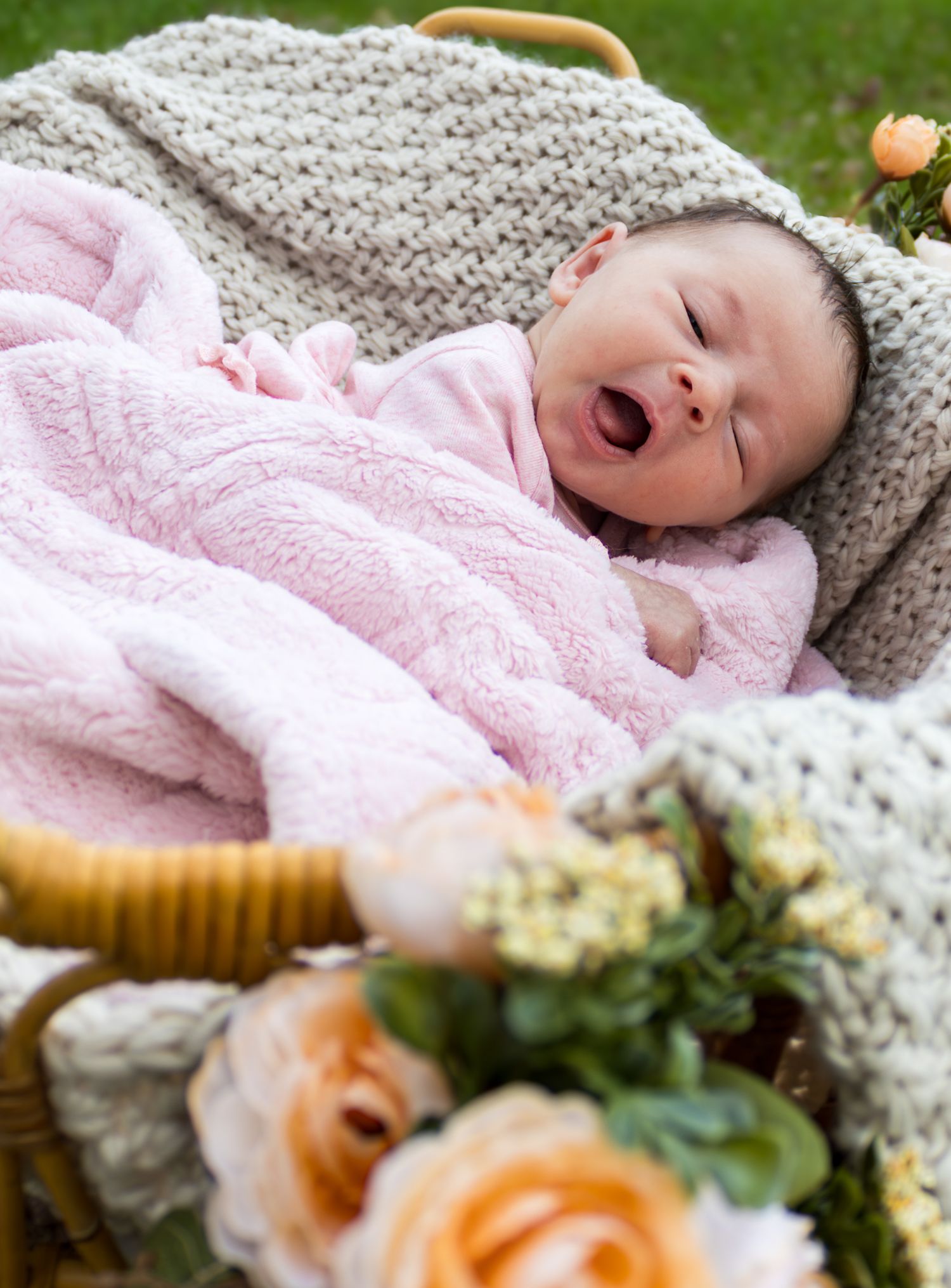 Newborn baby in pink outfit yawning in a wicker basket with gray knit blanket and flowers outdoors.
