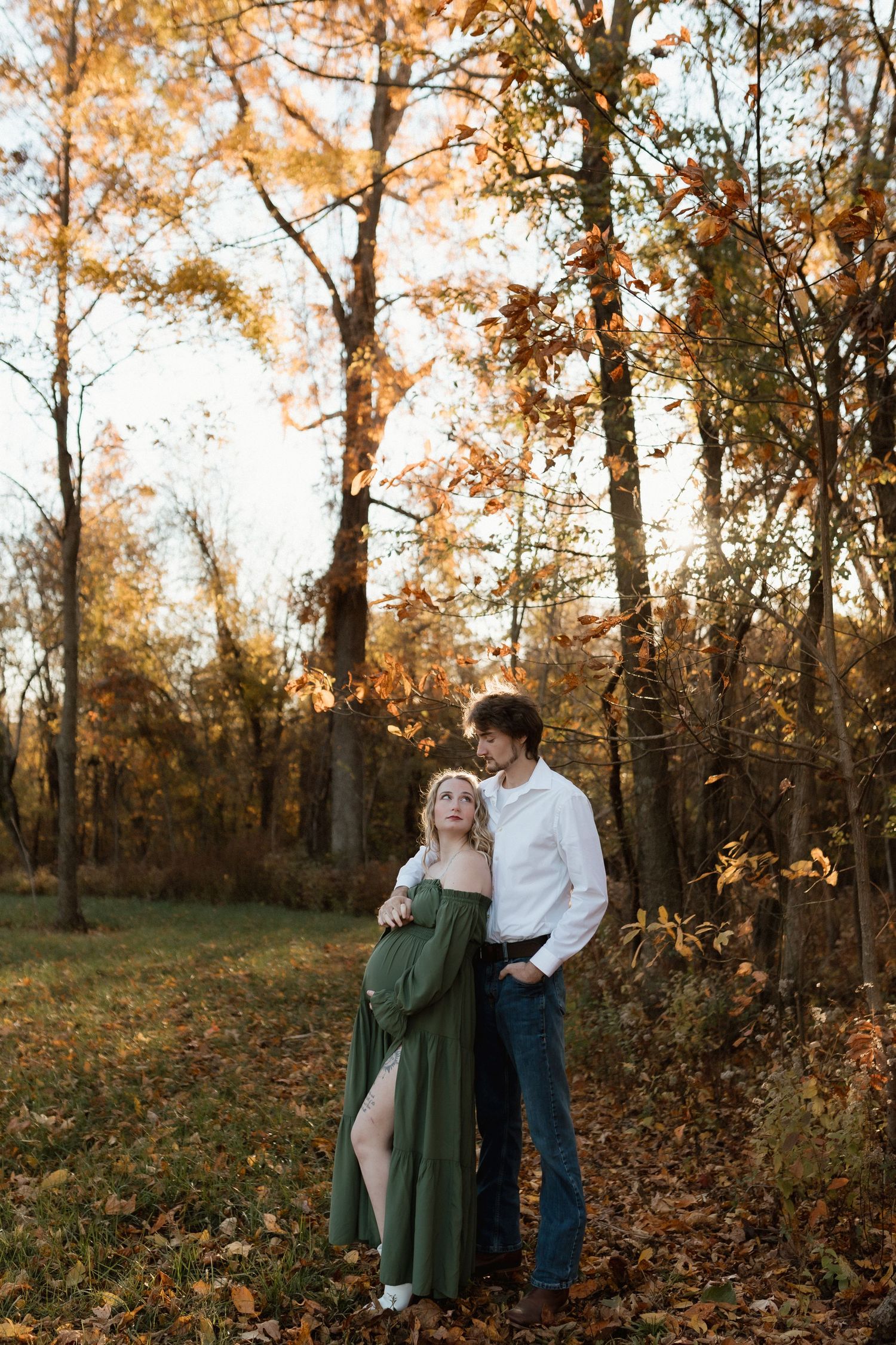 Couple embracing in autumn forest, pregnant woman in green dress with partner in white shirt among fall foliage.