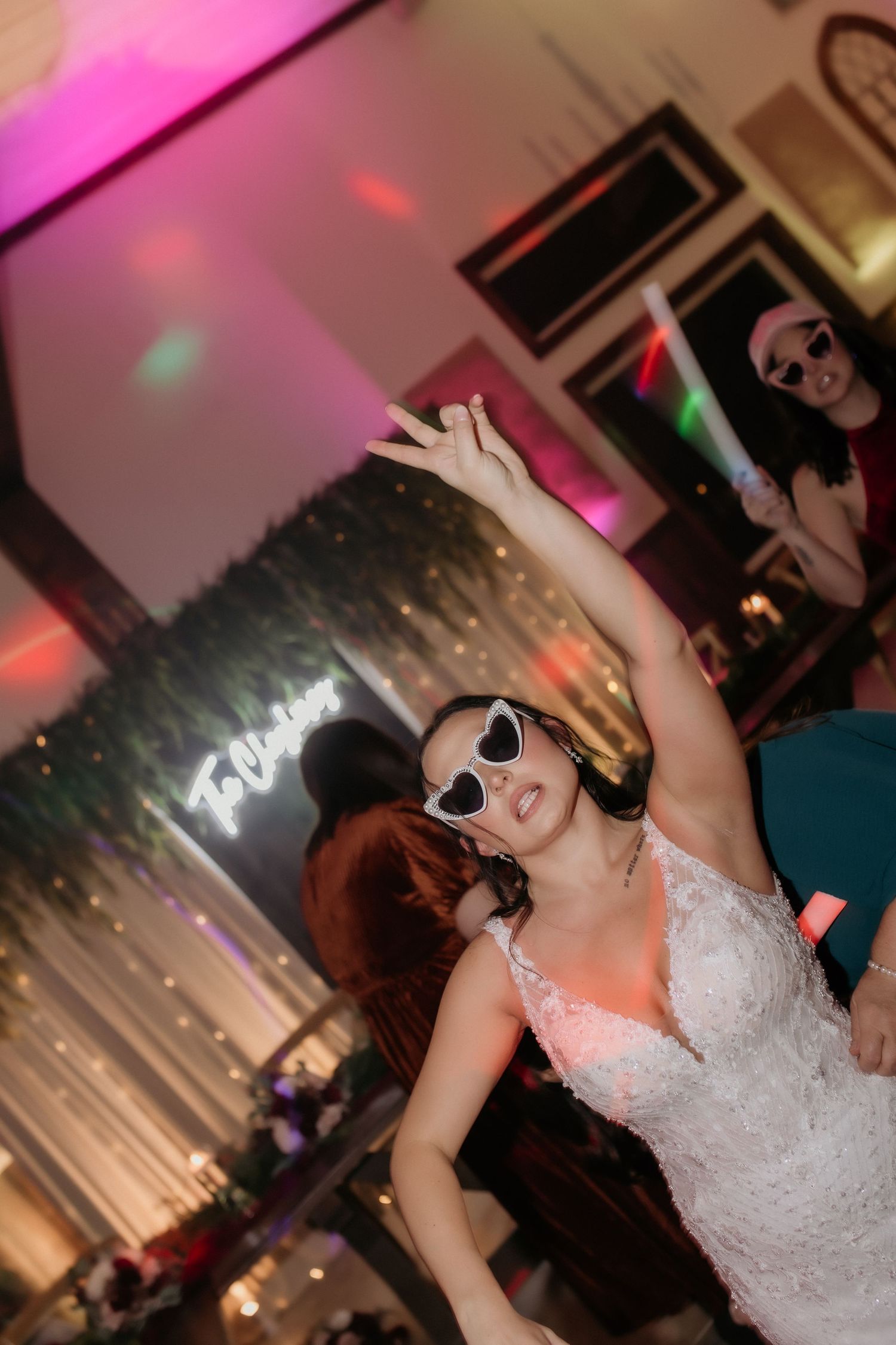 Bride in white lace gown dancing with arms raised wearing heart-shaped sunglasses at a vibrant wedding reception.