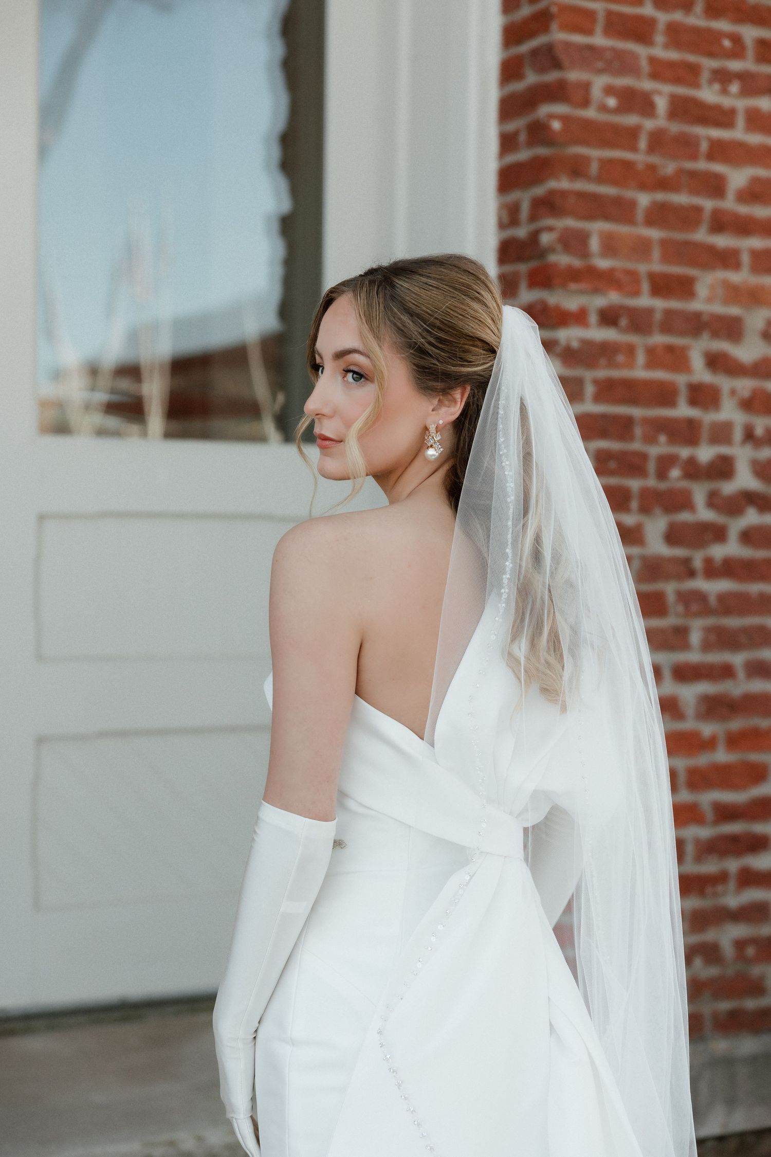 Bride in backless white wedding gown with veil and gloves glances back near a brick building.