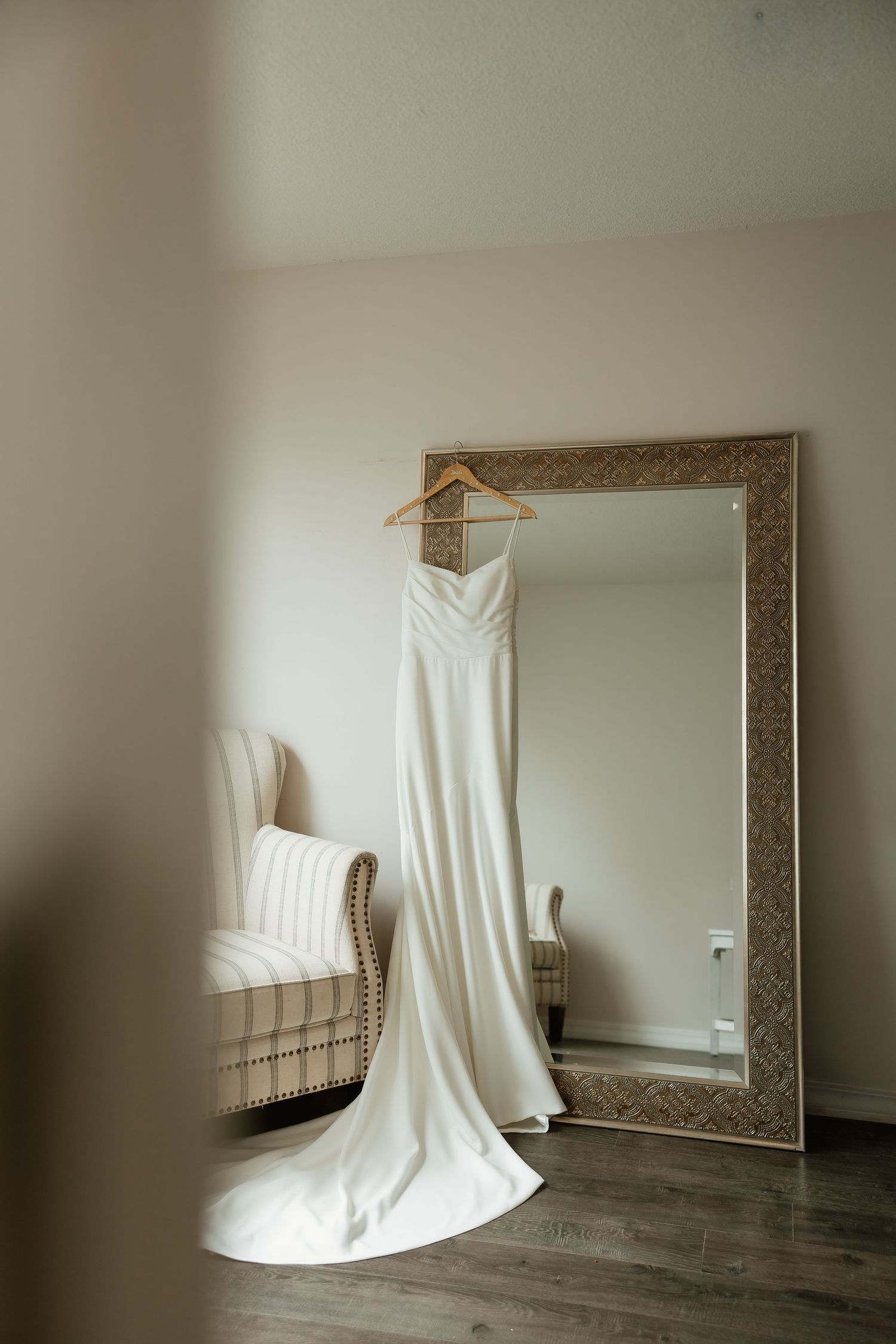 An elegant white wedding dress on a wooden hanger displayed against a large ornate floor mirror.