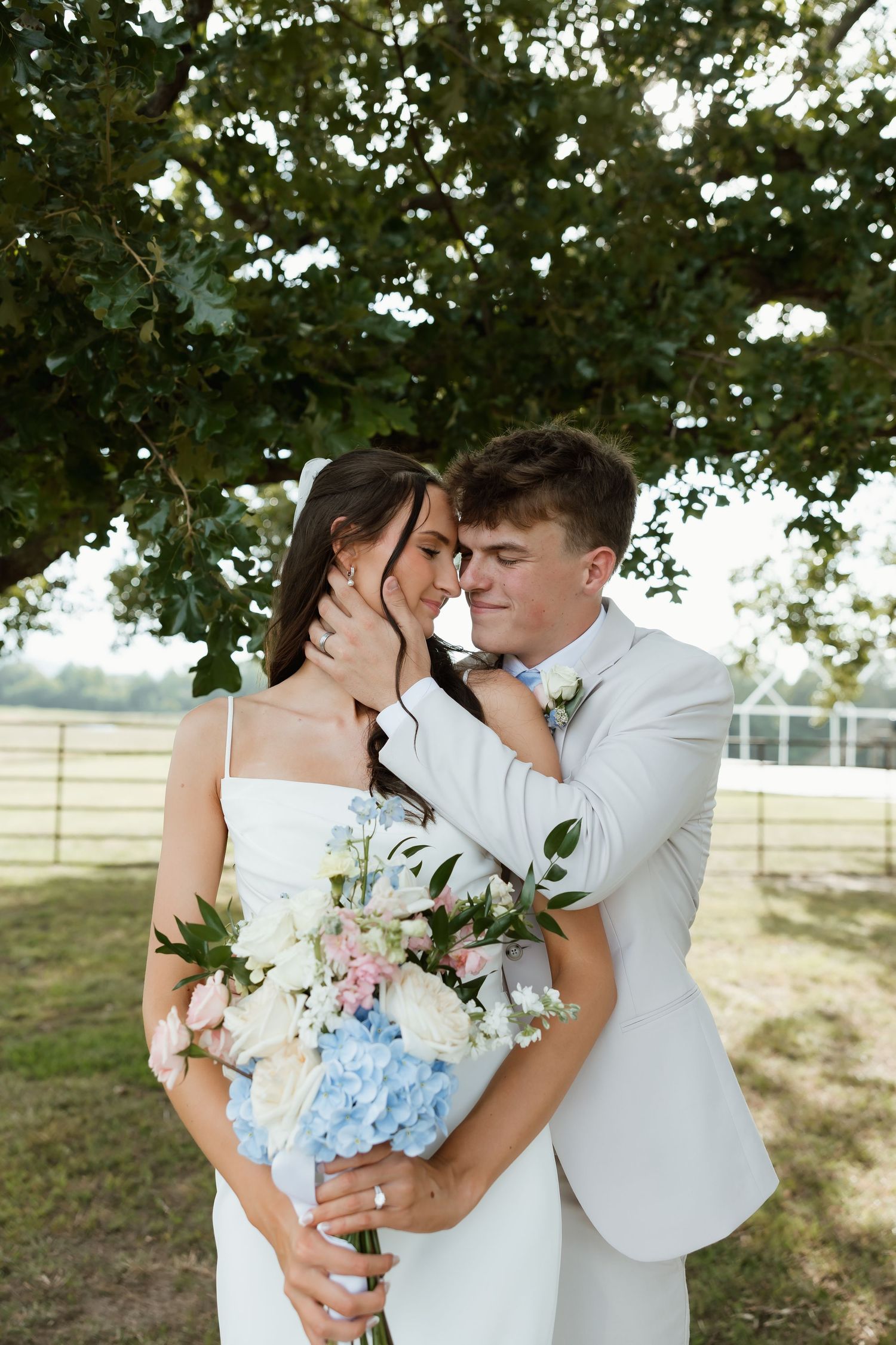 Groom tenderly holding bride's face as couple poses under oak tree with blue floral bouquet.