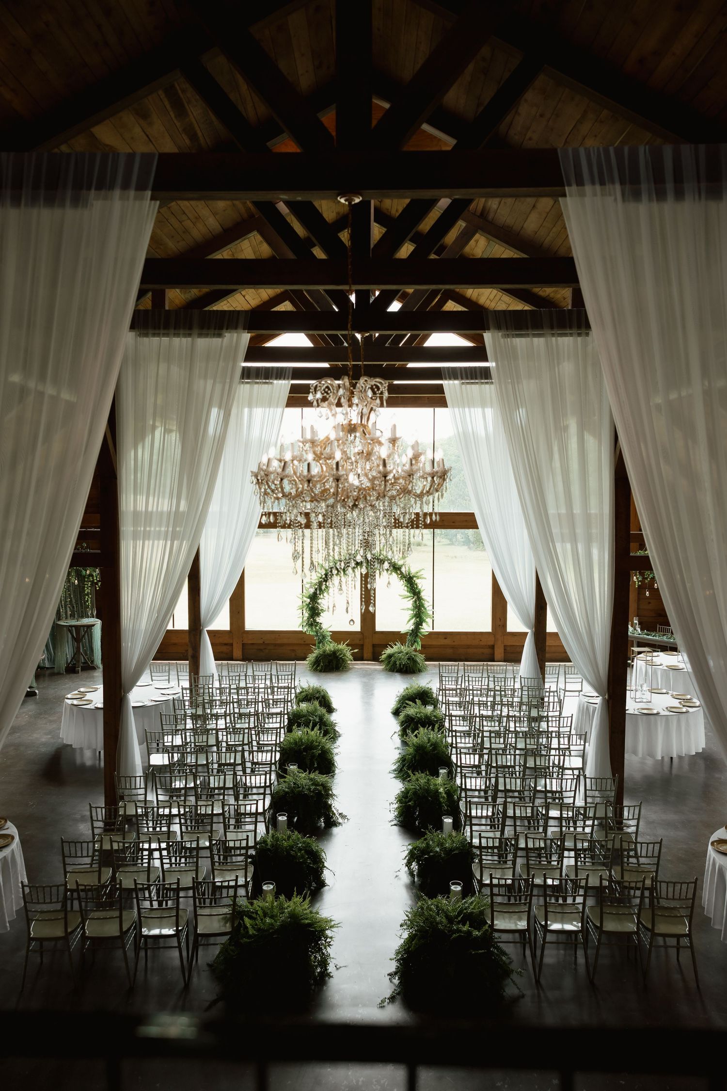Elegant barn wedding ceremony setup with chandelier, white drapes, greenery aisle, and chiavari chairs.