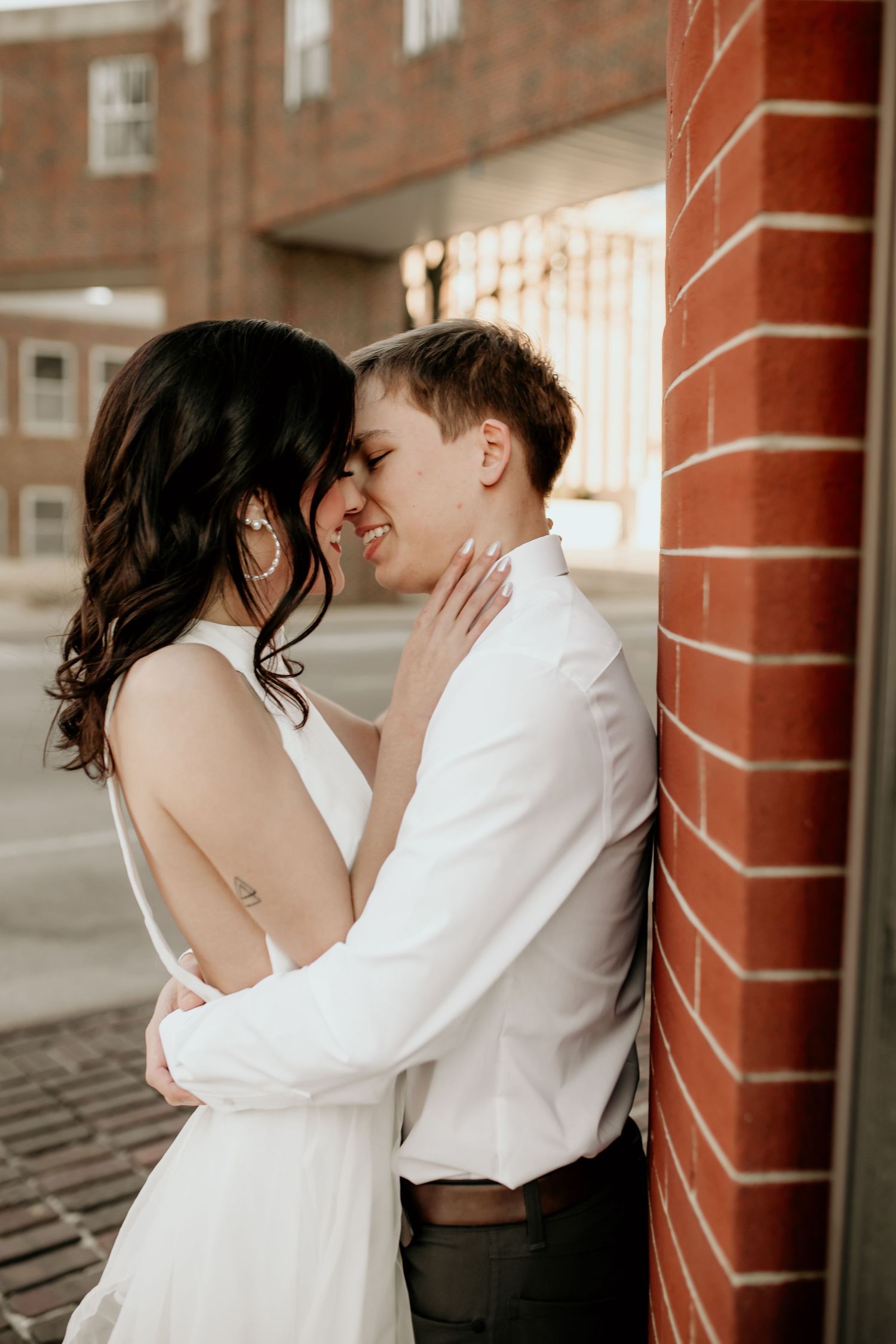 Couple sharing intimate almost-kiss moment leaning against brick wall in urban engagement photo session.