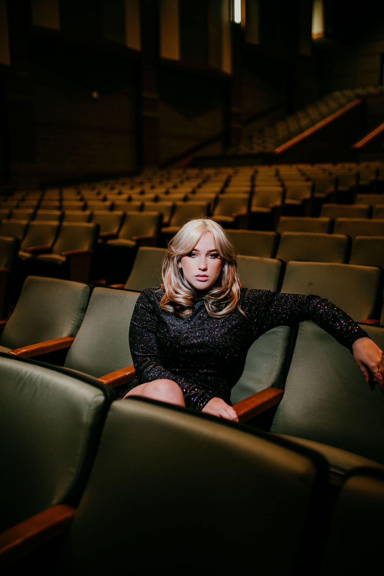 Blonde woman in black sparkle dress sitting in empty theater seats.