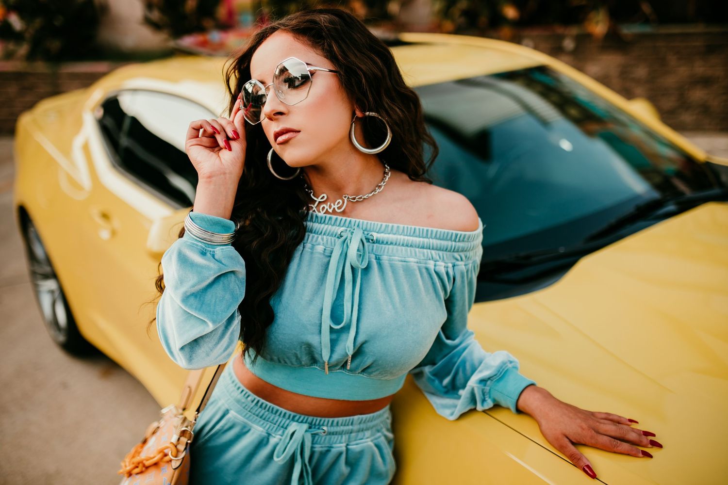 Stylish woman in blue crop set leaning on yellow sports car wearing sunglasses.