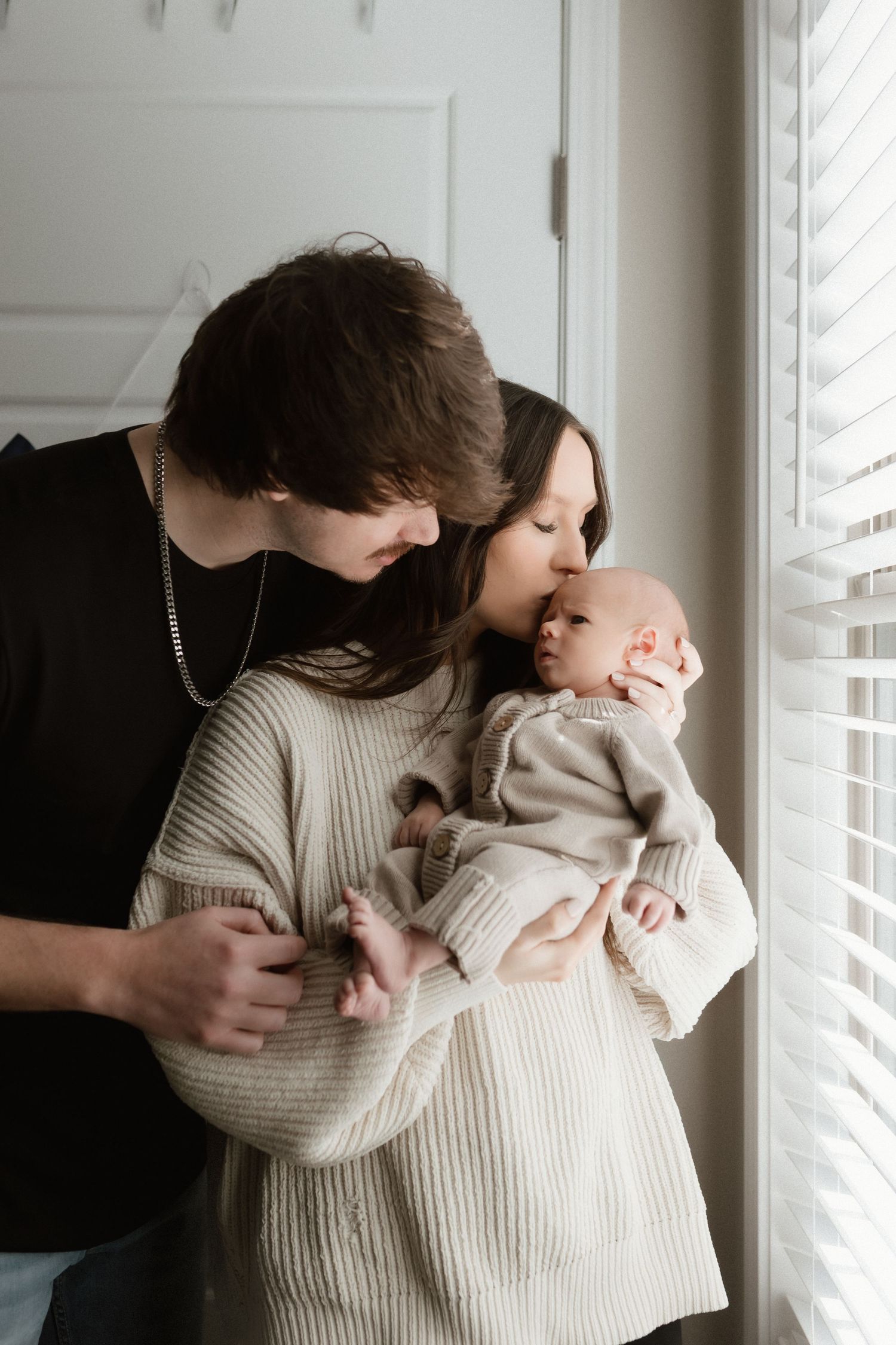 Young couple tenderly kissing newborn baby by window with soft natural light filtering through blinds.