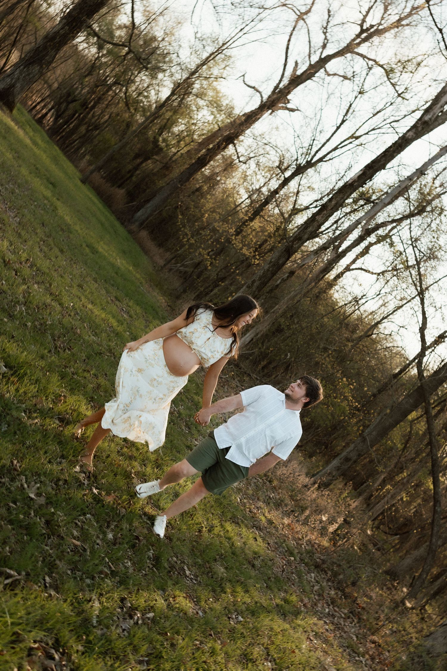 Pregnant woman in a floral white dress dancing with her partner in a sunlit wooded field during golden hour.