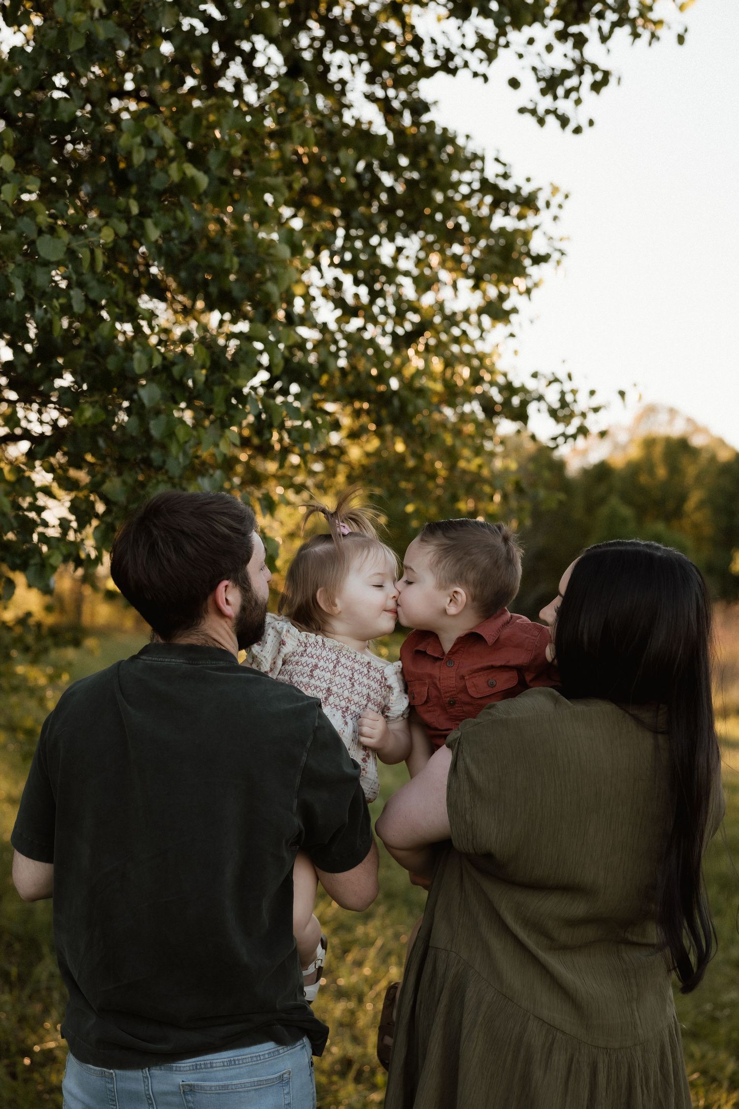 Parents holding two young children who share a sweet kiss under golden sunlit trees outdoors.