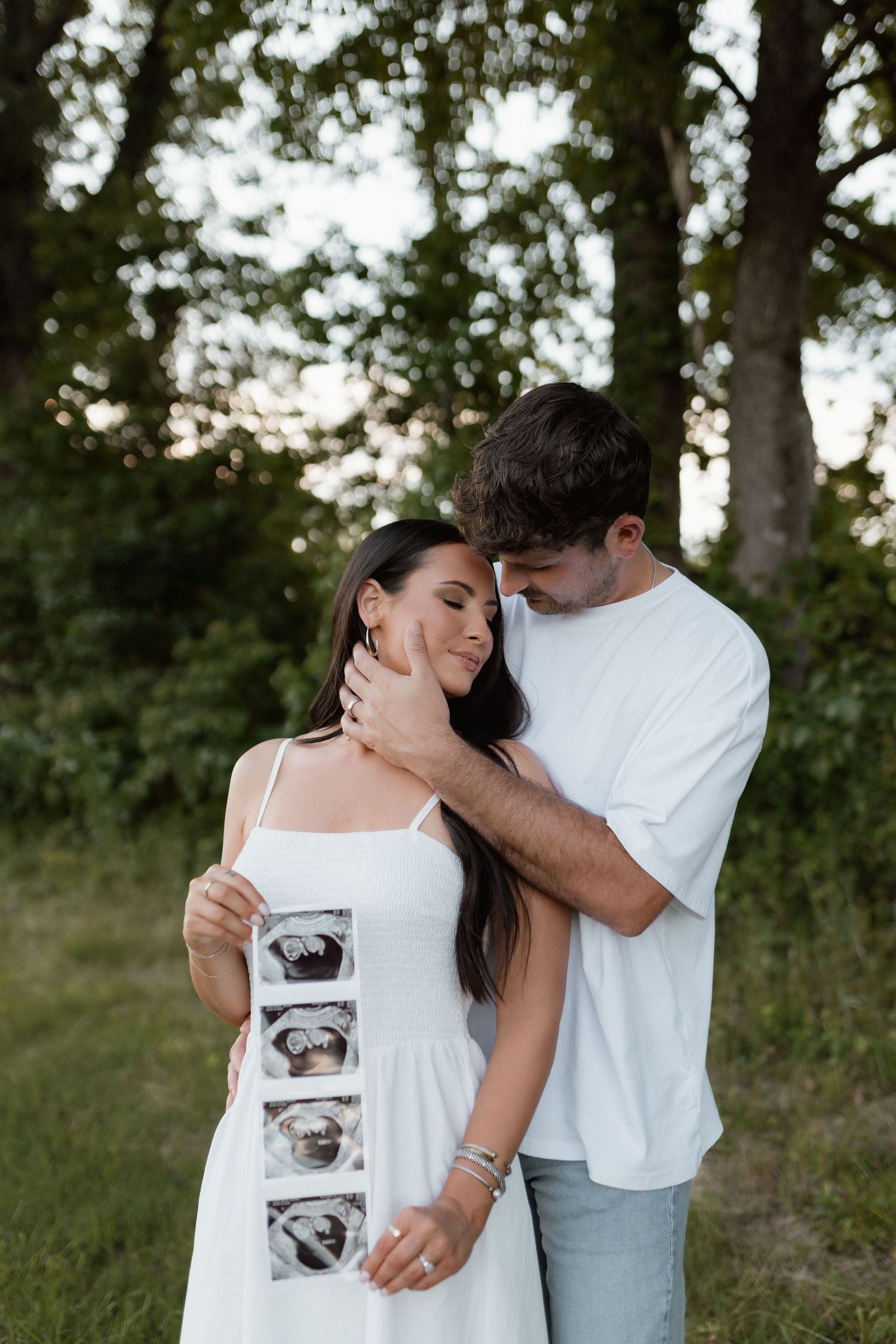 Couple embracing outdoors while woman holds ultrasound photos announcing their pregnancy near green trees.