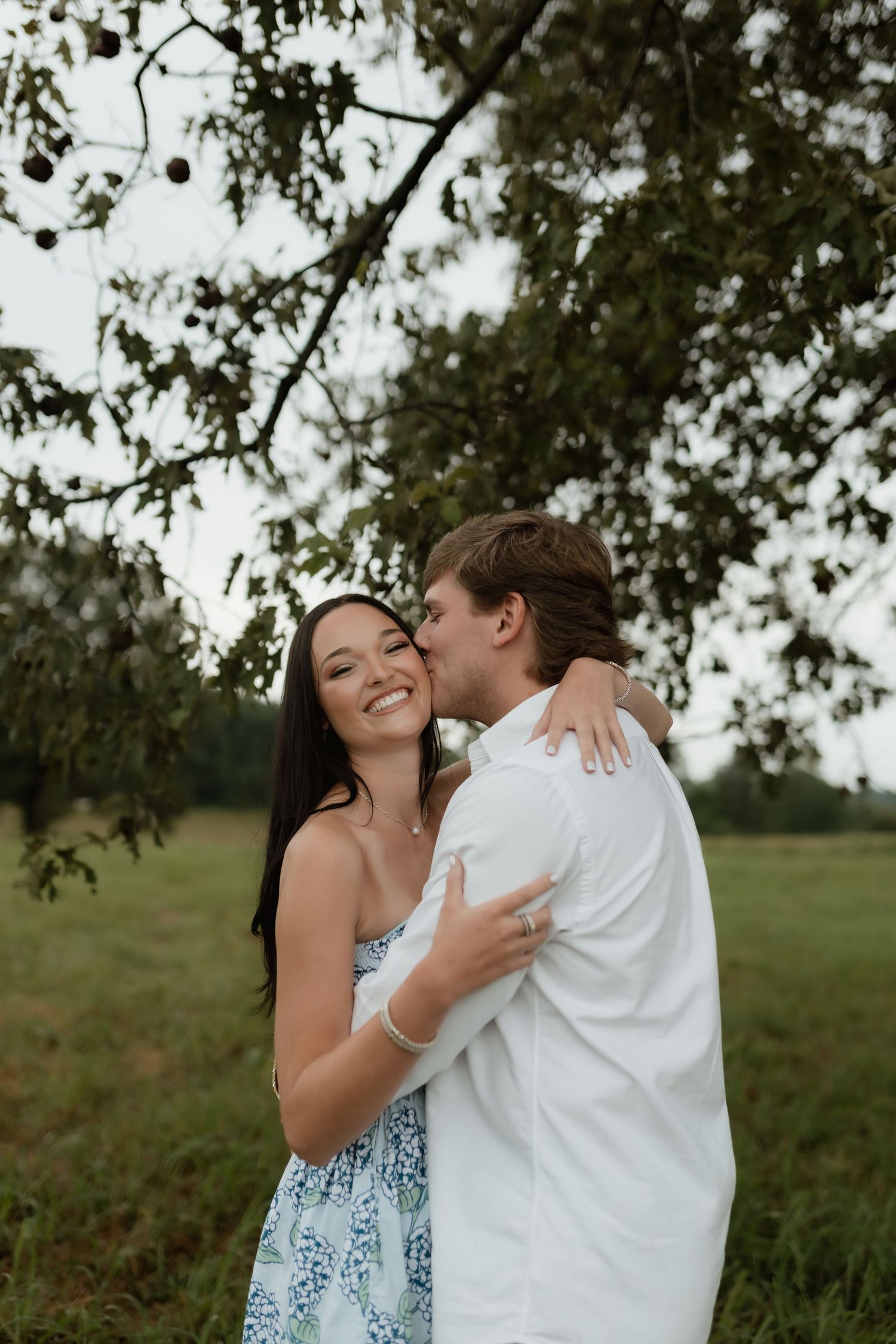 A smiling woman in a floral dress embraces a man in a white shirt who kisses her cheek outdoors.