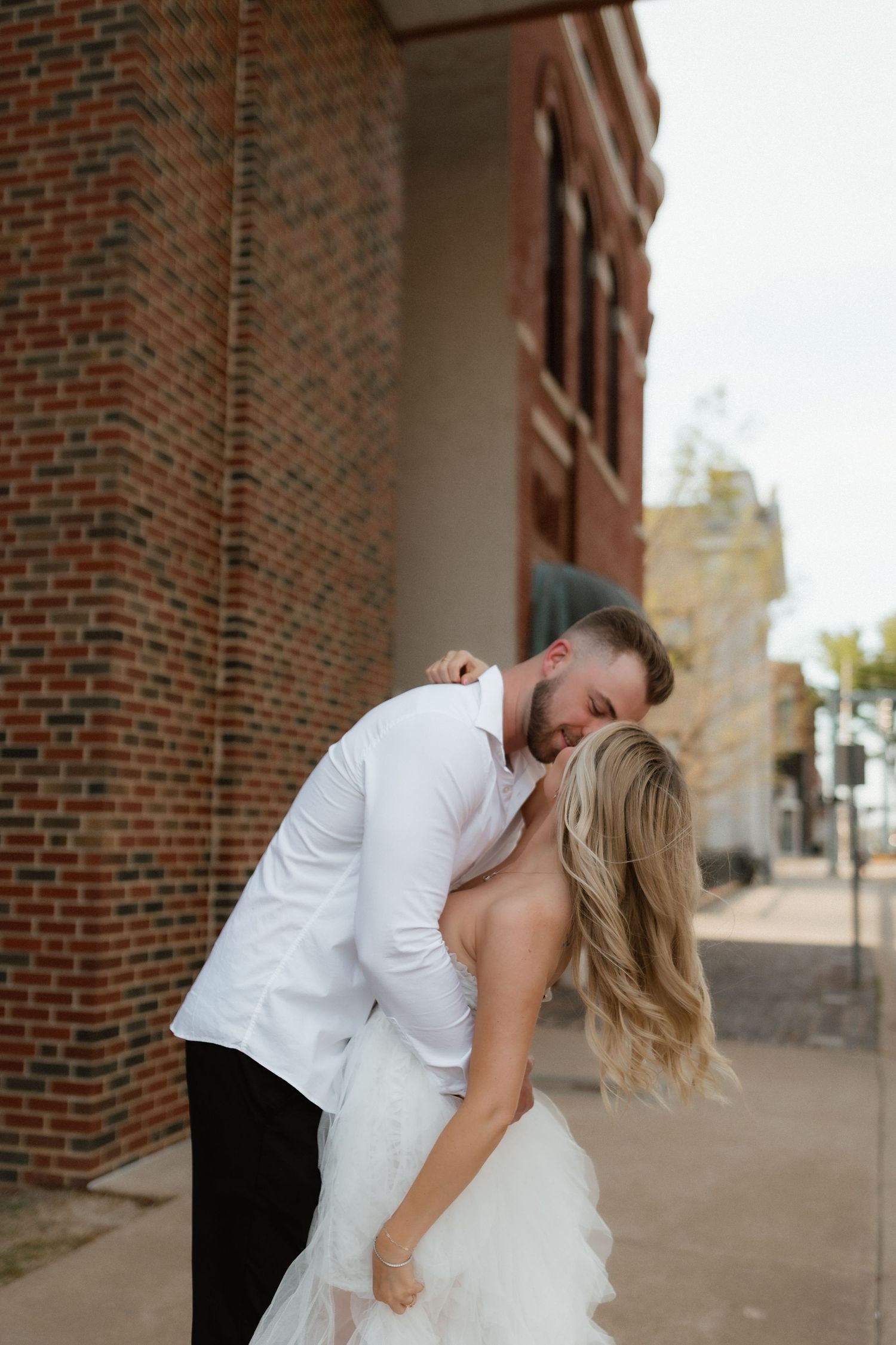 Groom in white shirt dipping blonde bride in flowing white dress outside brick building on wedding day.