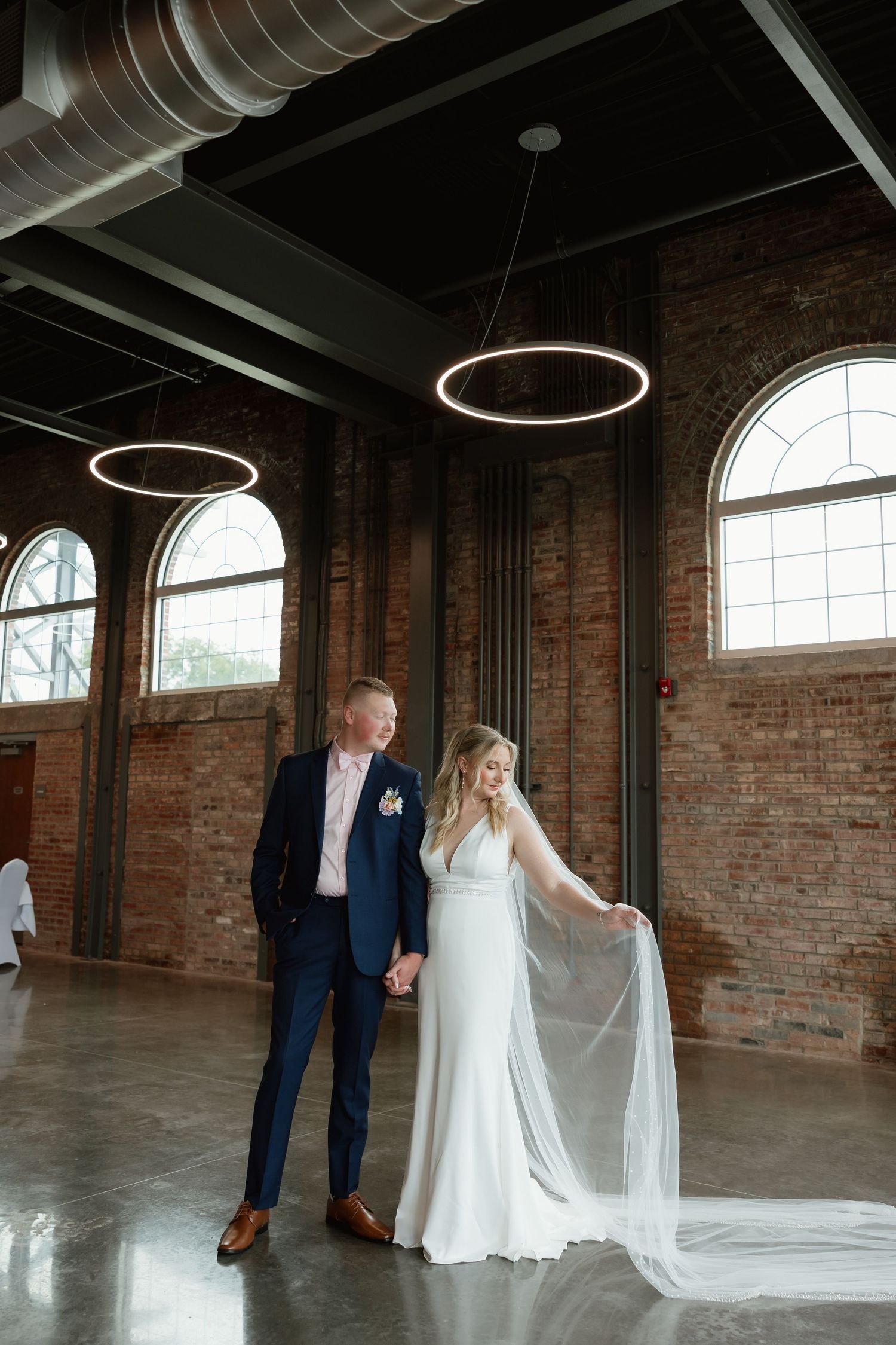 Bride and groom hold hands in industrial chic brick venue as bride elegantly holds her flowing cathedral veil.