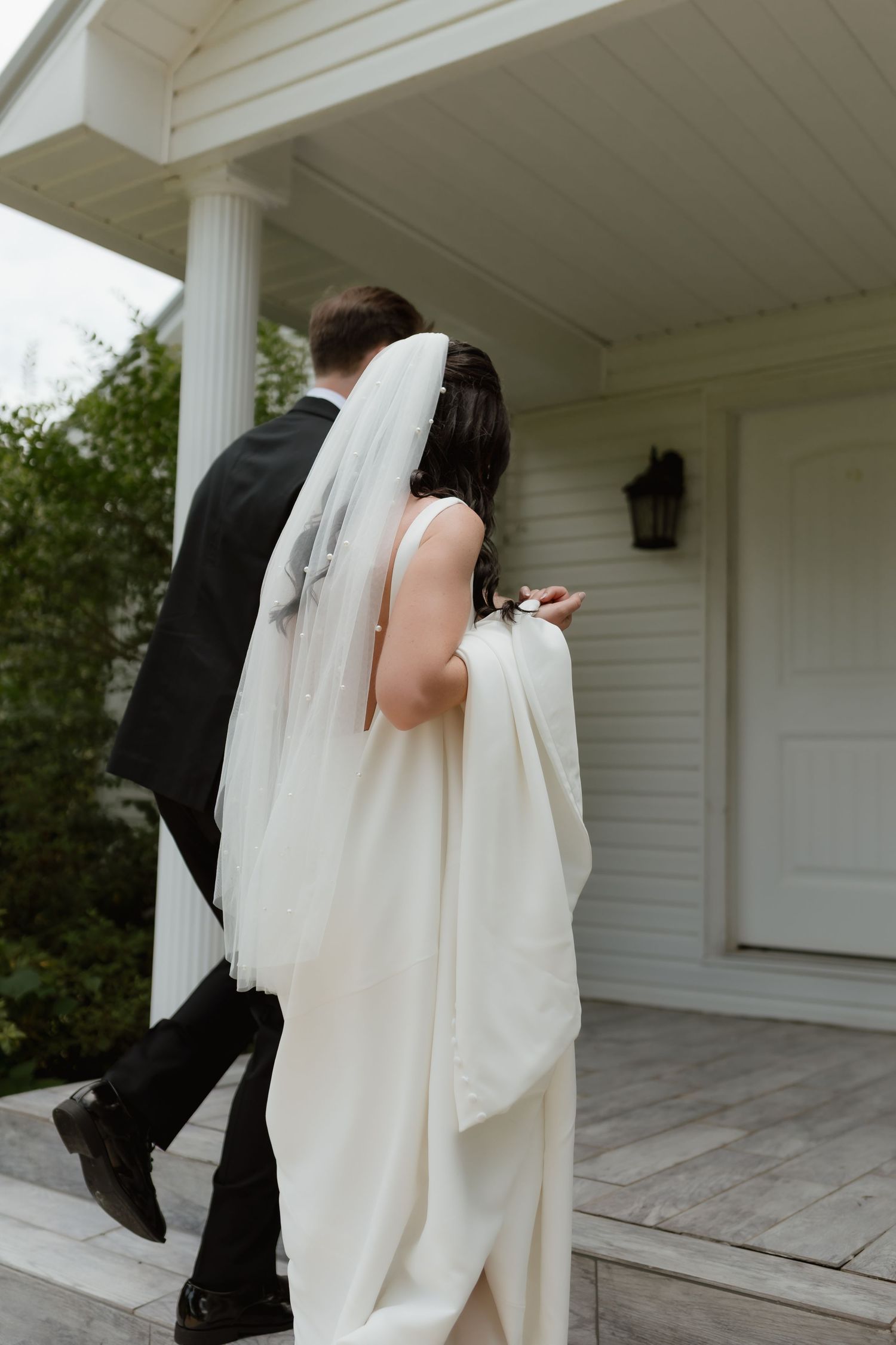 Bride and groom walking up porch steps of a white colonial-style building on wedding day.