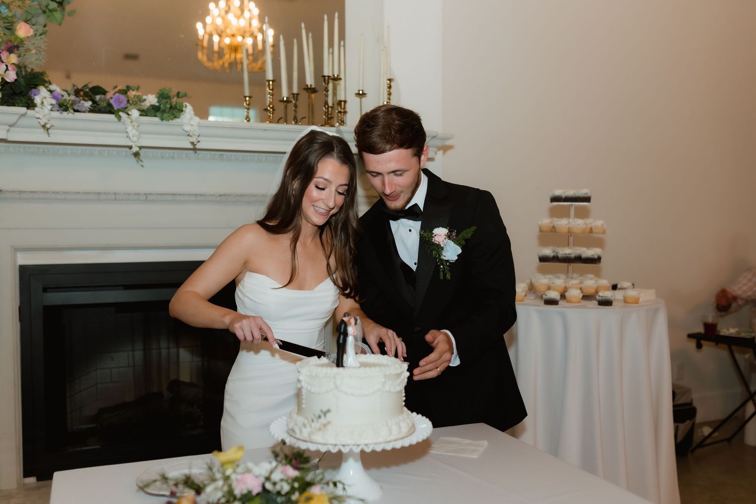 Bride and groom cutting white wedding cake near fireplace at reception.