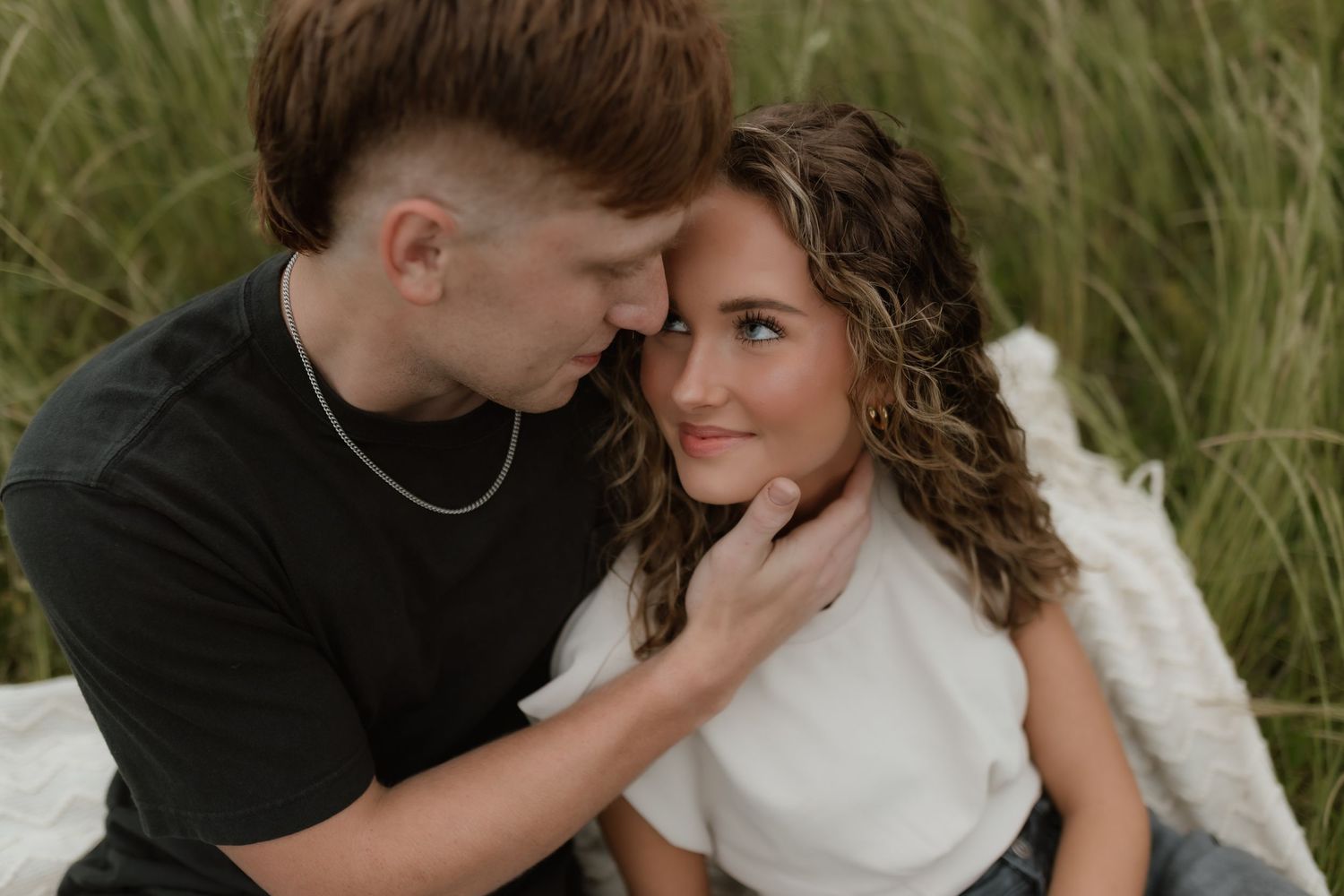 Young couple sitting in tall grass, man tenderly holding woman's face as they lean close together.