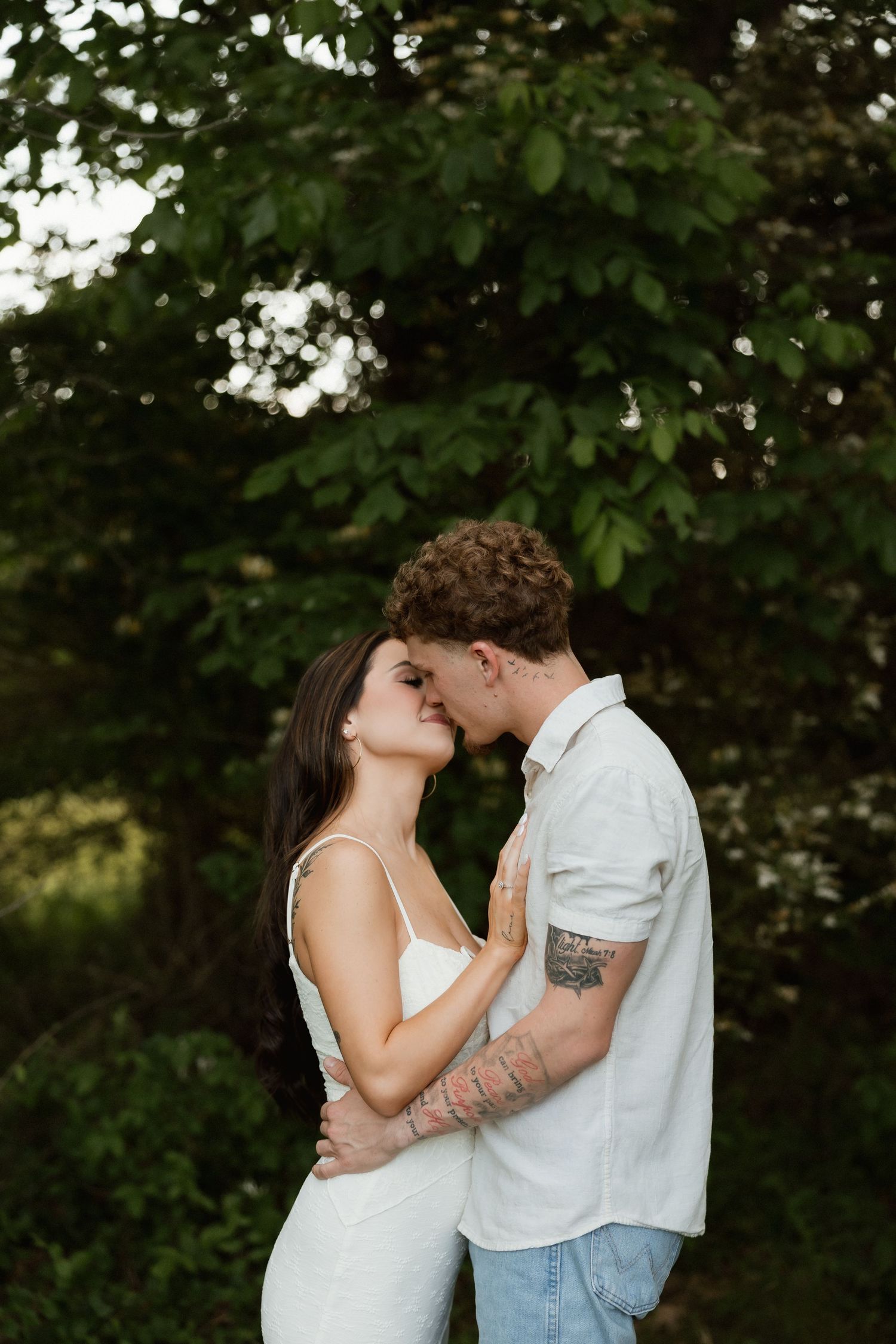 A tattooed couple shares a kiss surrounded by lush green trees in natural light.