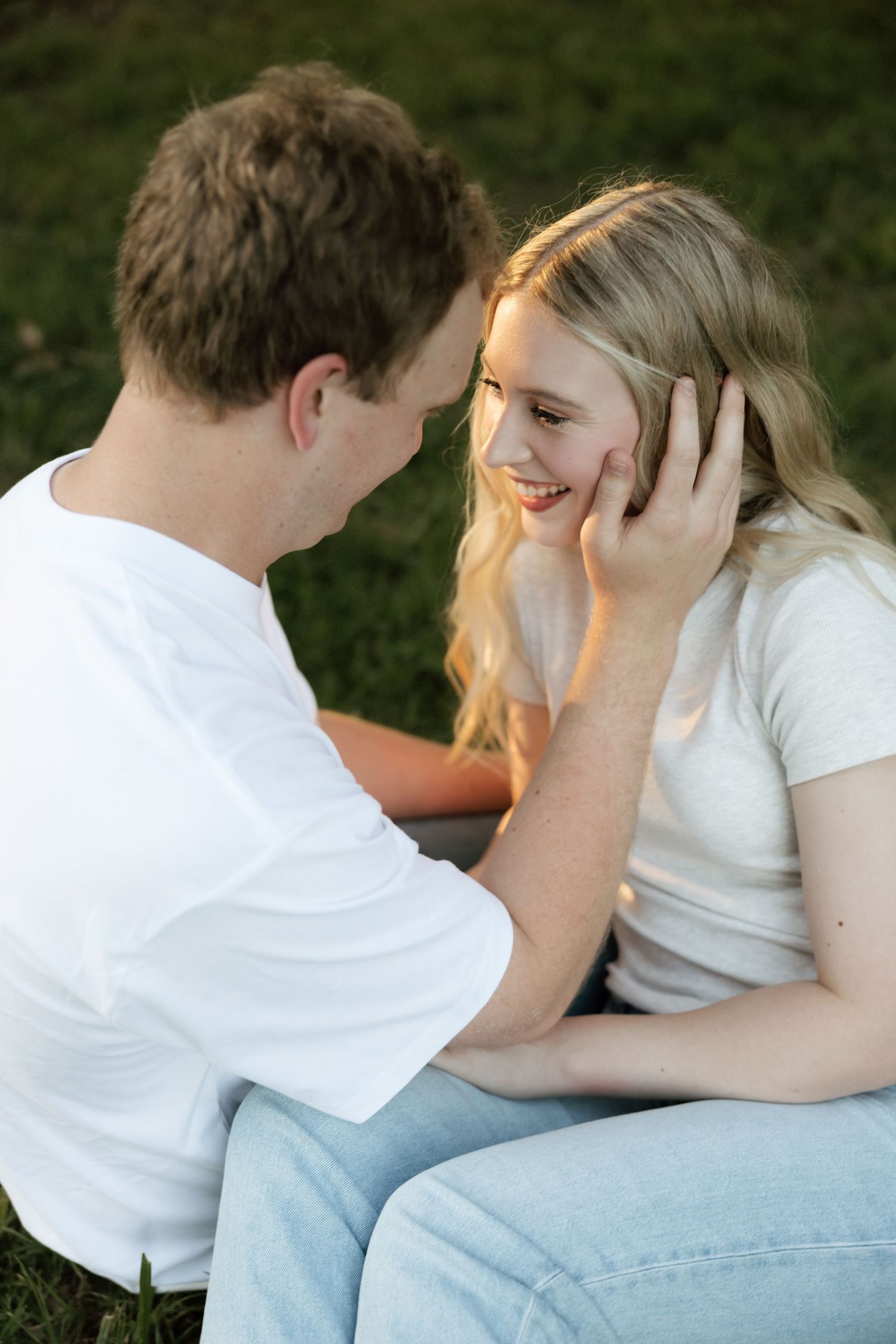 A man gently touches a smiling blonde woman's face as they sit together on grass in warm sunlight.