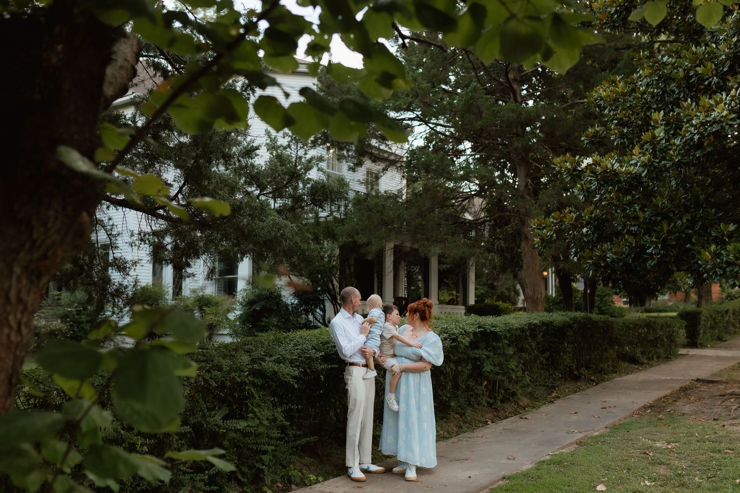 Family posing together on a garden path surrounded by lush trees and hedges near a white historic home.
