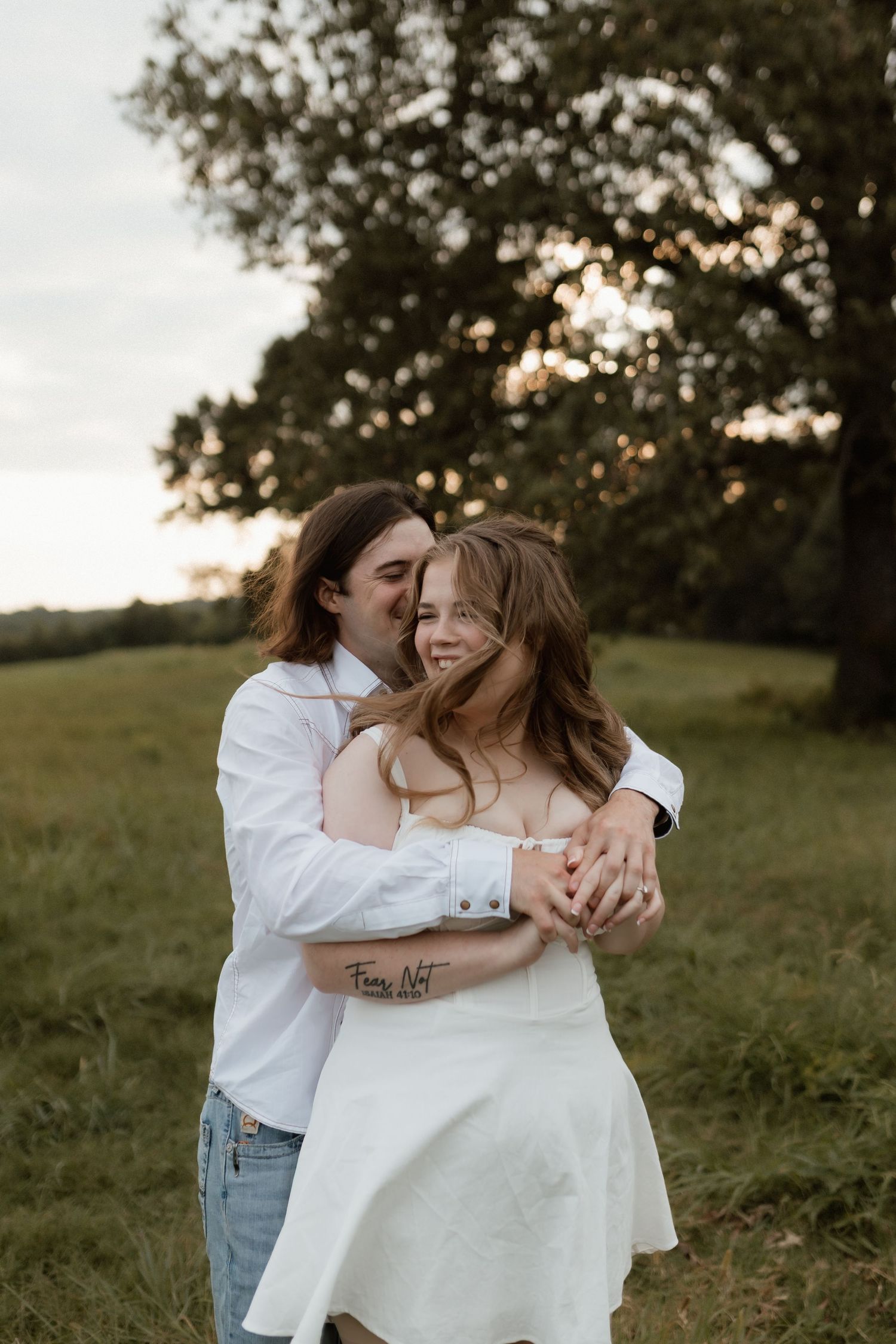 Couple embracing at sunset in green field, woman in white dress laughing as man holds her from behind near large tree.