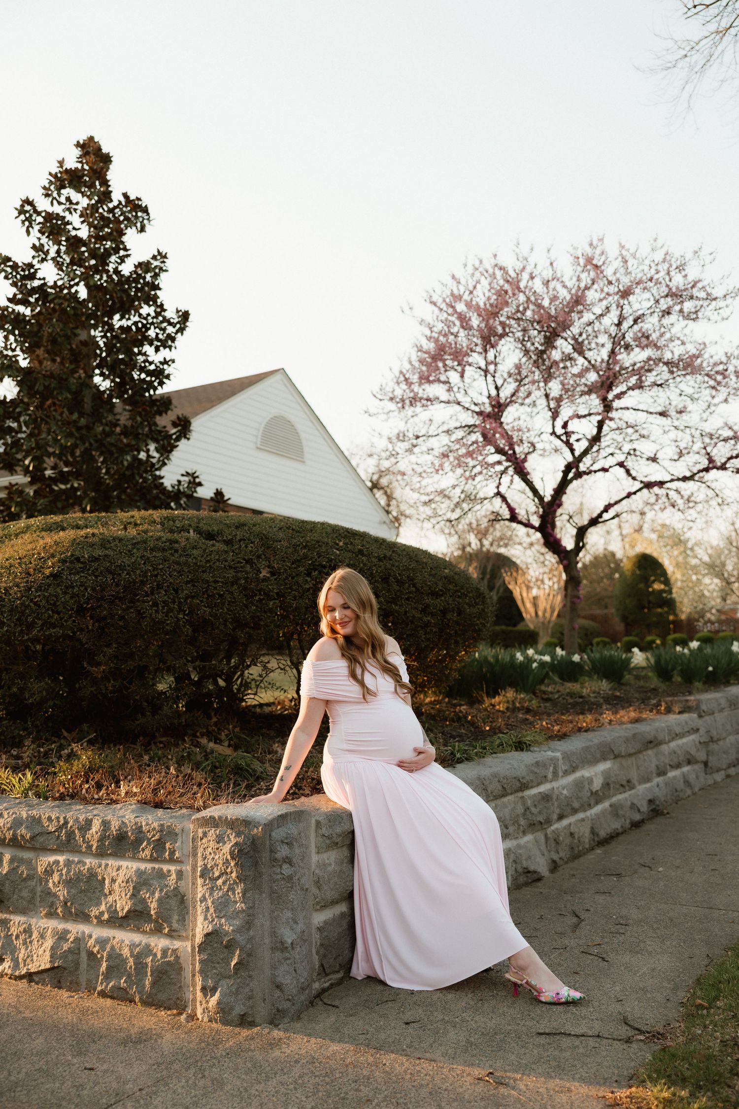 Pregnant woman in pink maxi dress sitting on stone wall surrounded by spring garden blooms.
