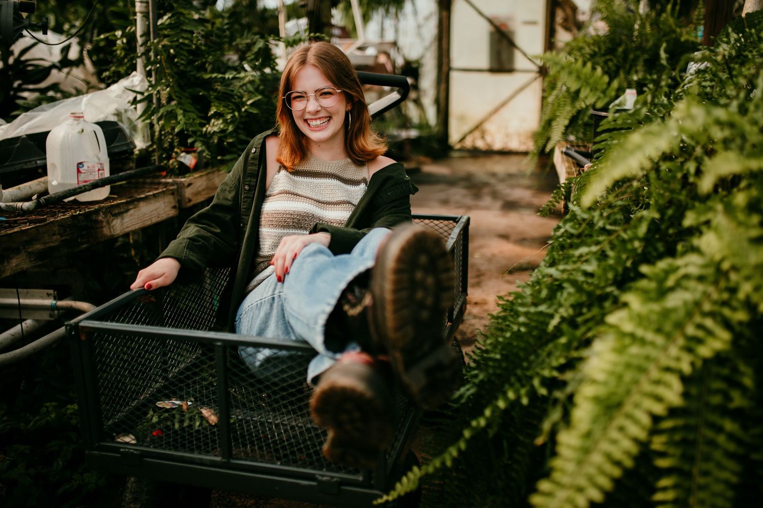 Cheerful redhead teen girl laughing while sitting in a metal cart surrounded by greenhouse plants.