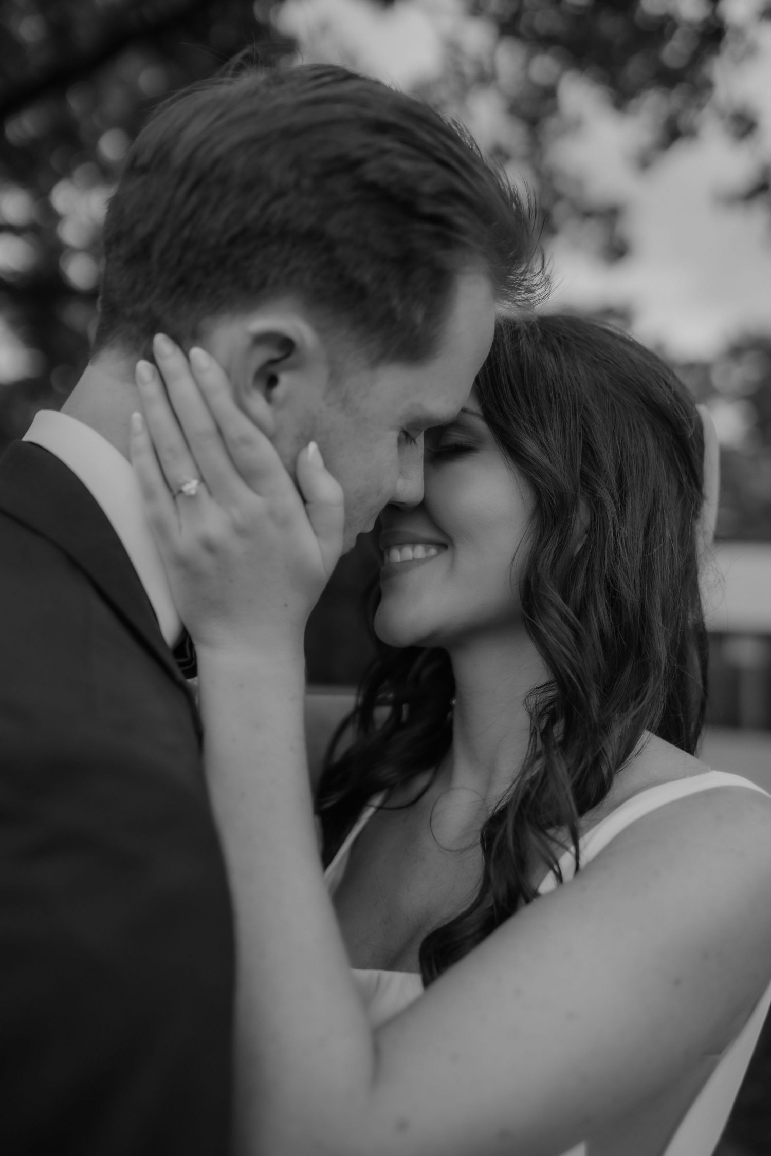 Black and white photo of smiling bride holding grooms face as they lean in to kiss.