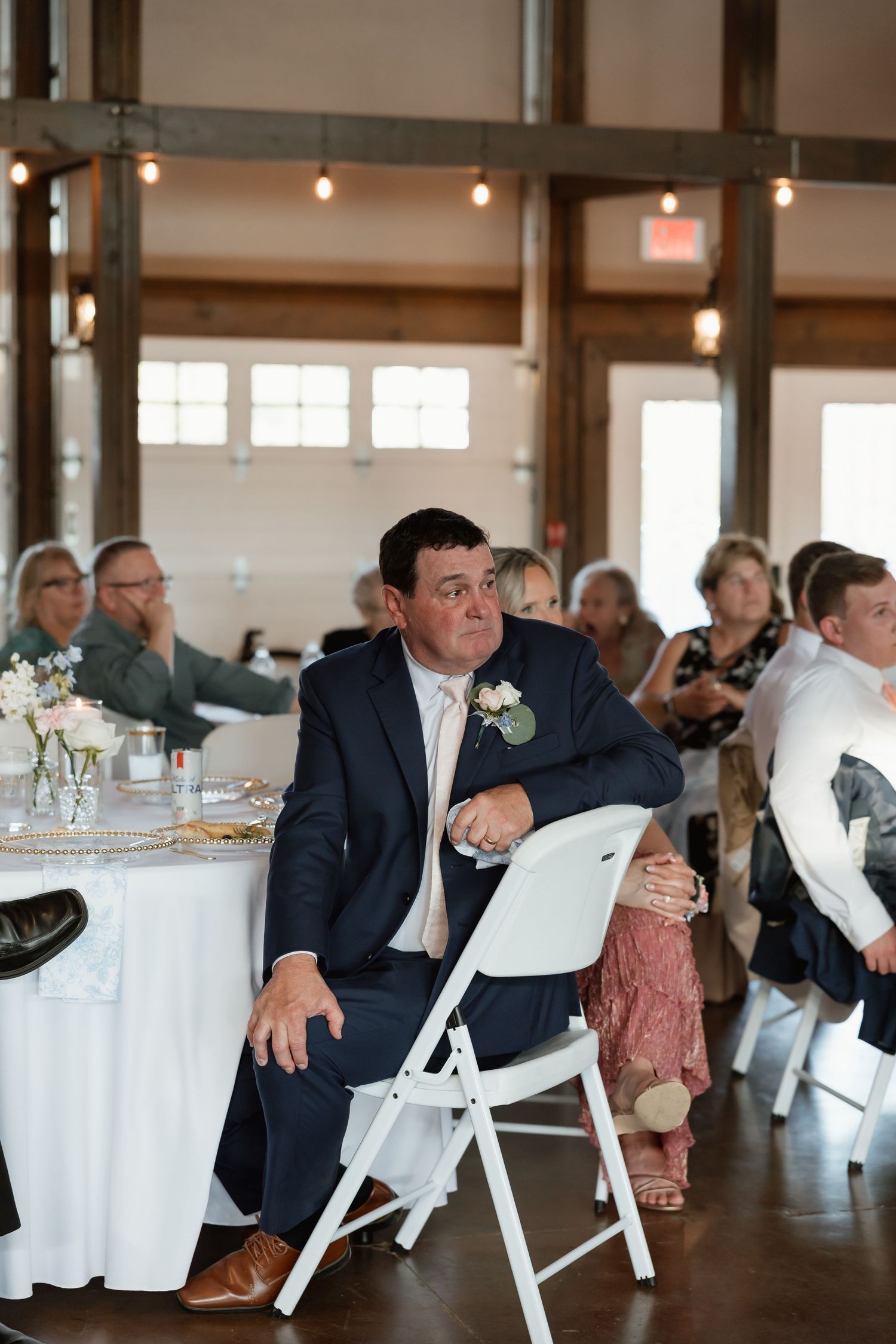 Groom in navy suit with boutonniere seated at wedding reception watching emotional moment.