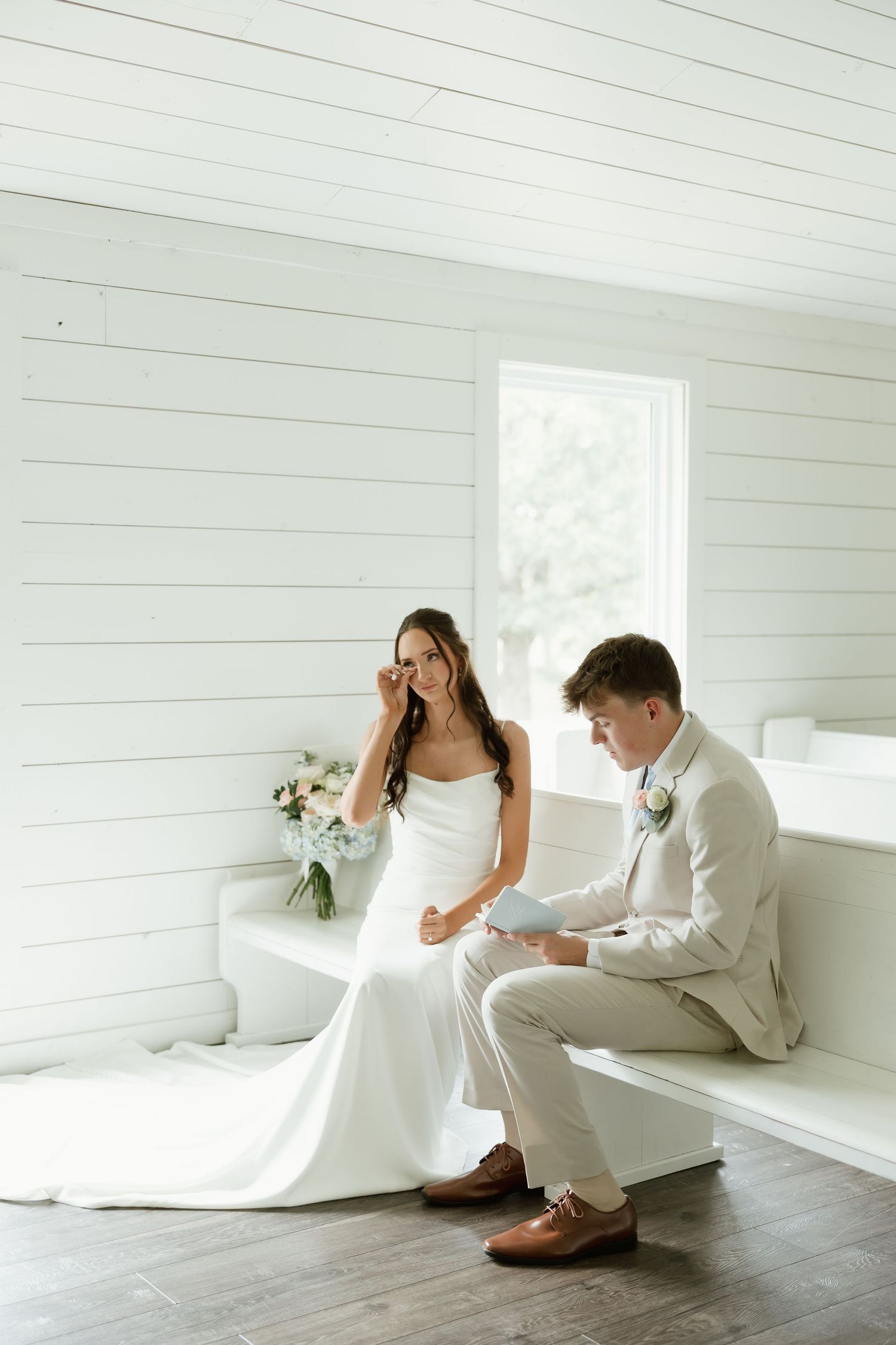 A bride wipes a tear while a groom reads vows on a white bench in a bright shiplap room.