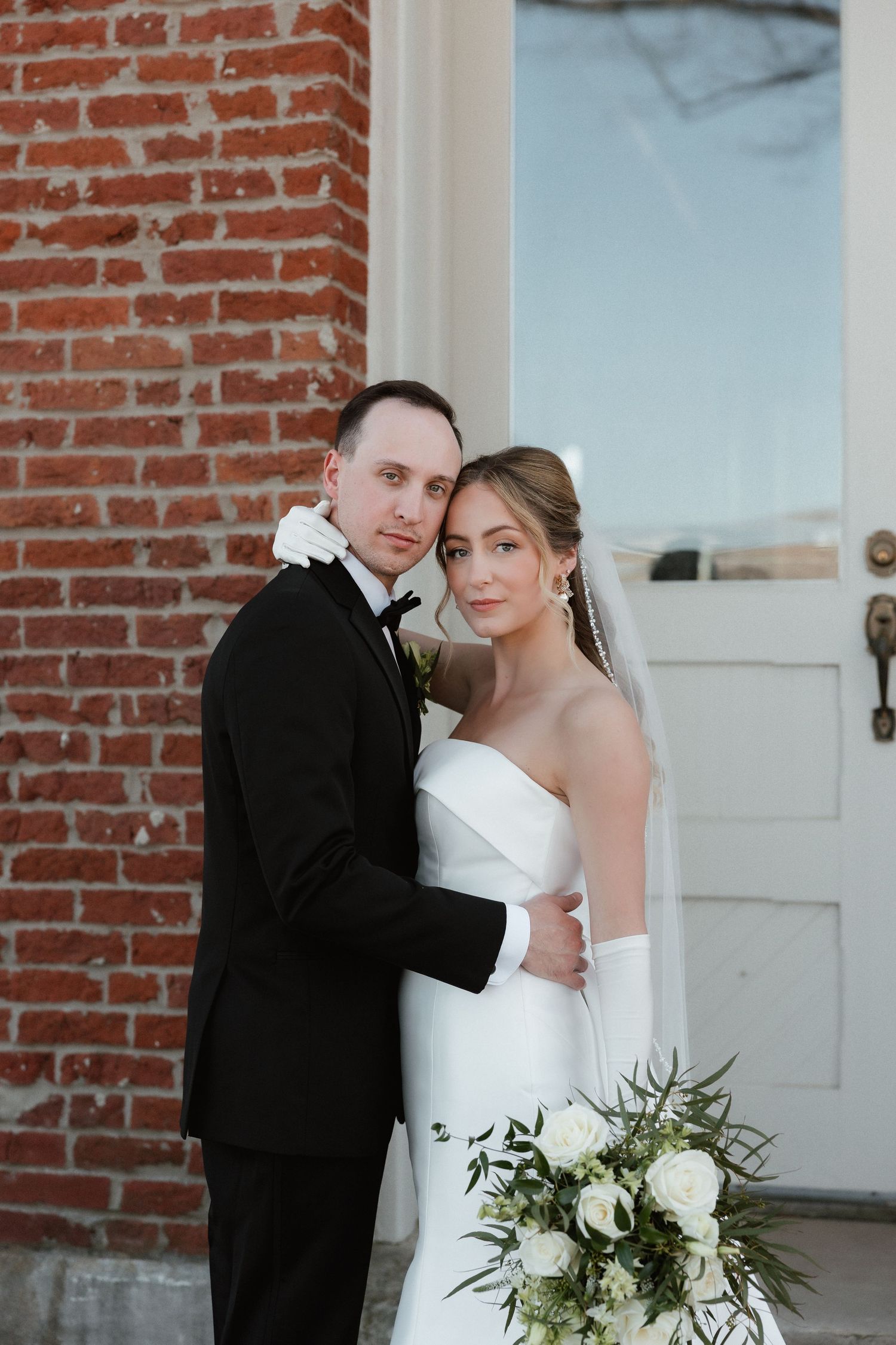 Bride and groom embrace tenderly holding white floral bouquet outside a rustic brick building.