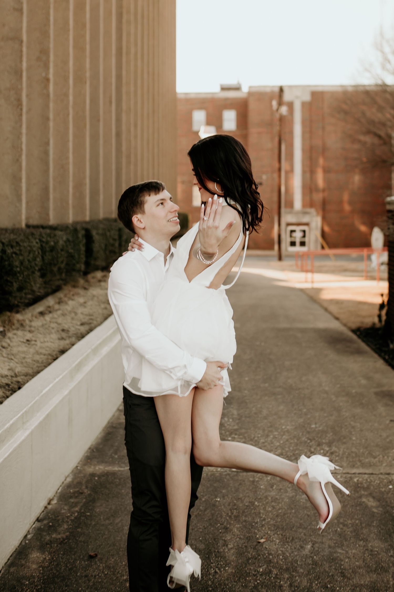 Man lifting woman in white mini dress and heels while she shows engagement ring in urban setting.