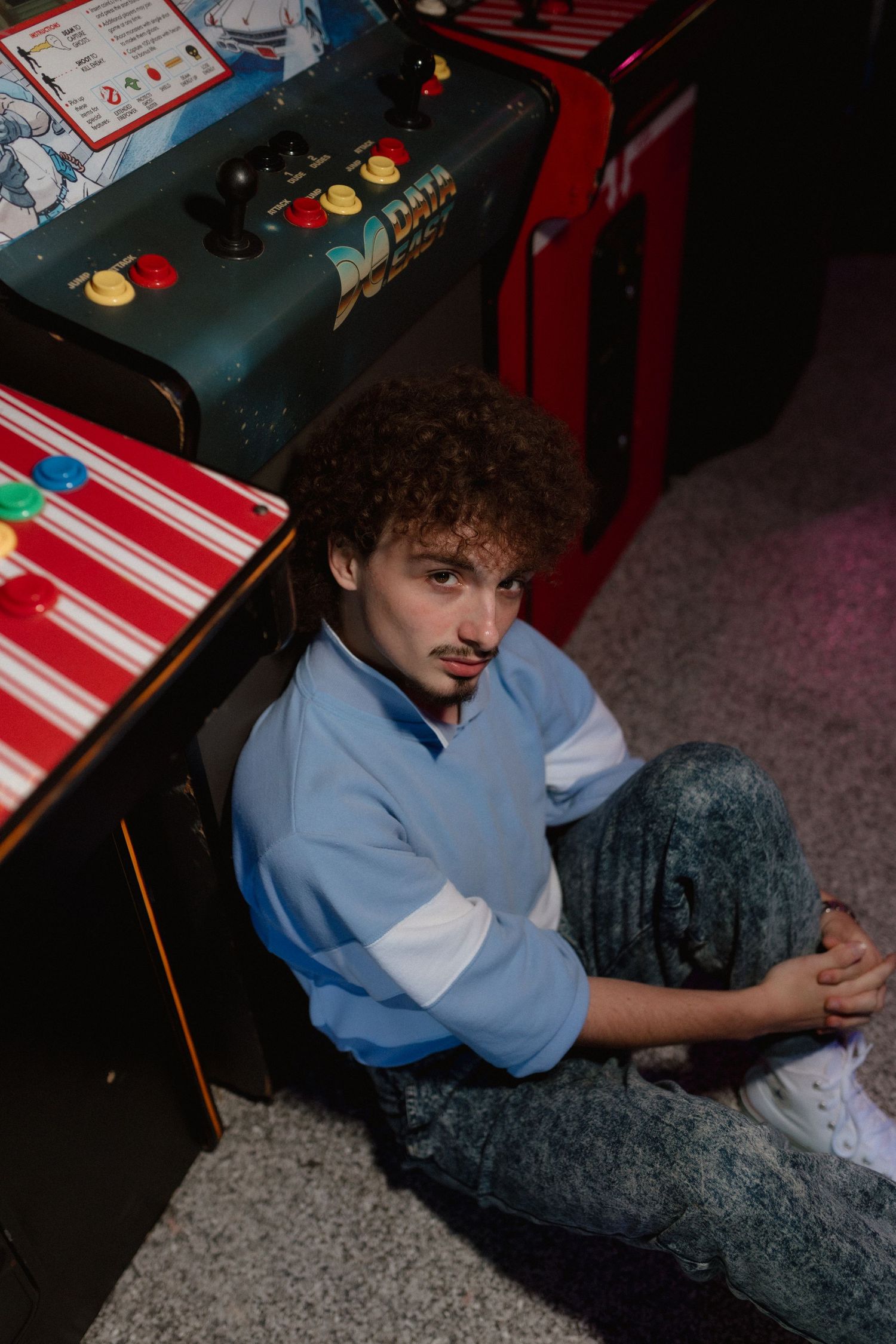 Young man with curly hair sitting on arcade floor in blue retro sweatshirt and acid wash jeans.