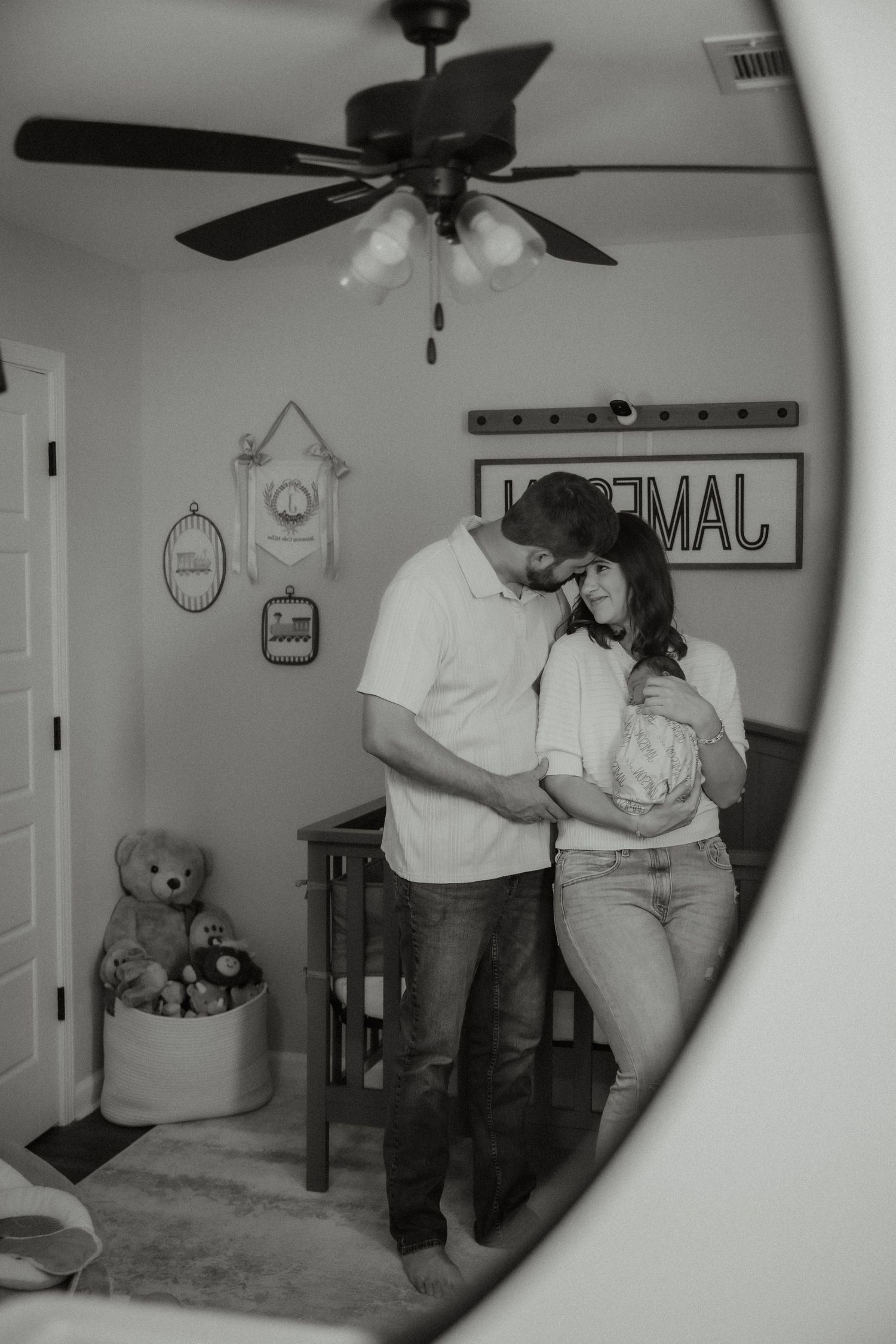 Black and white mirror reflection of couple embracing in decorated nursery with crib and teddy bear.