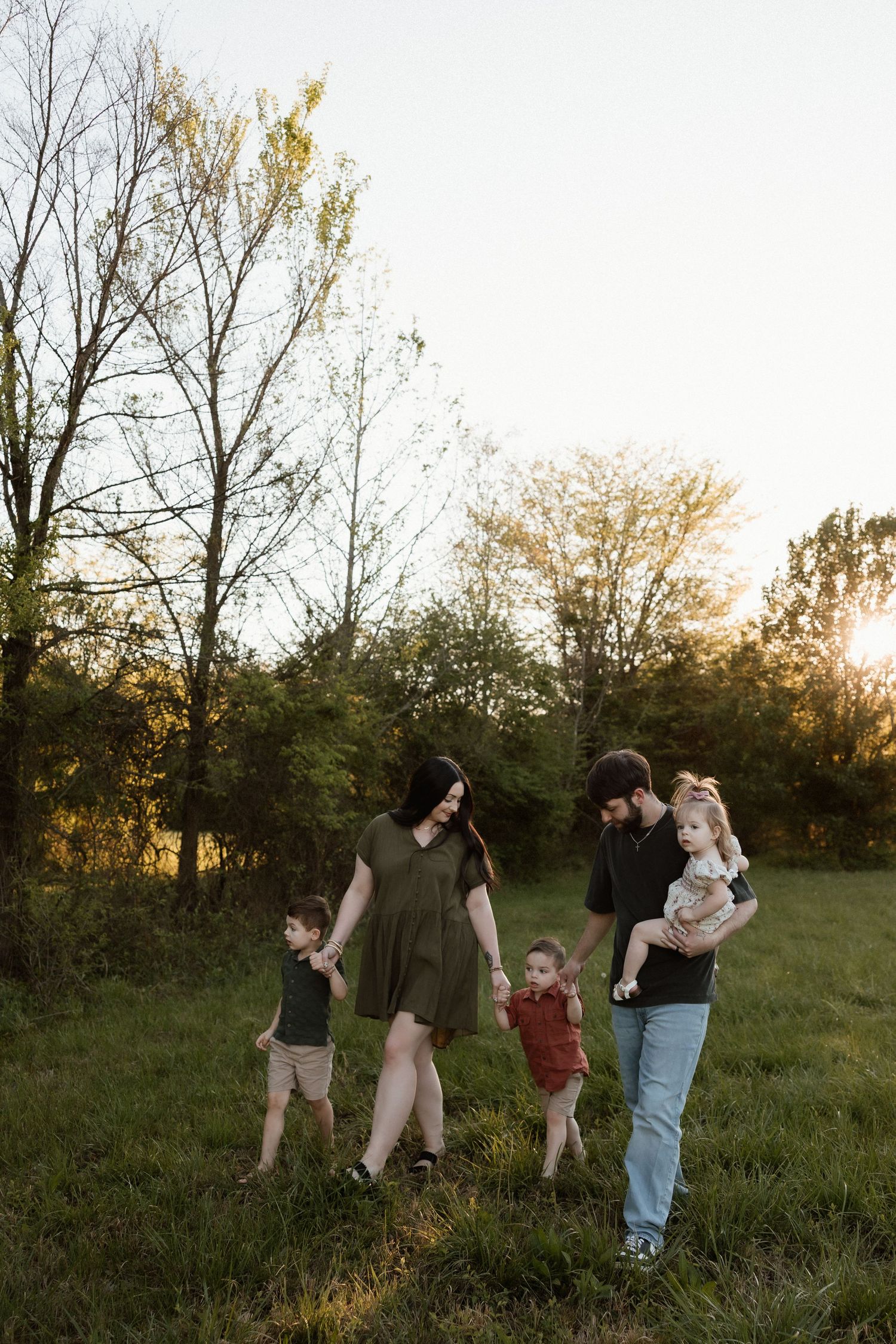 Family of five walking through a sunlit green field at golden hour, parents holding hands with three young children.