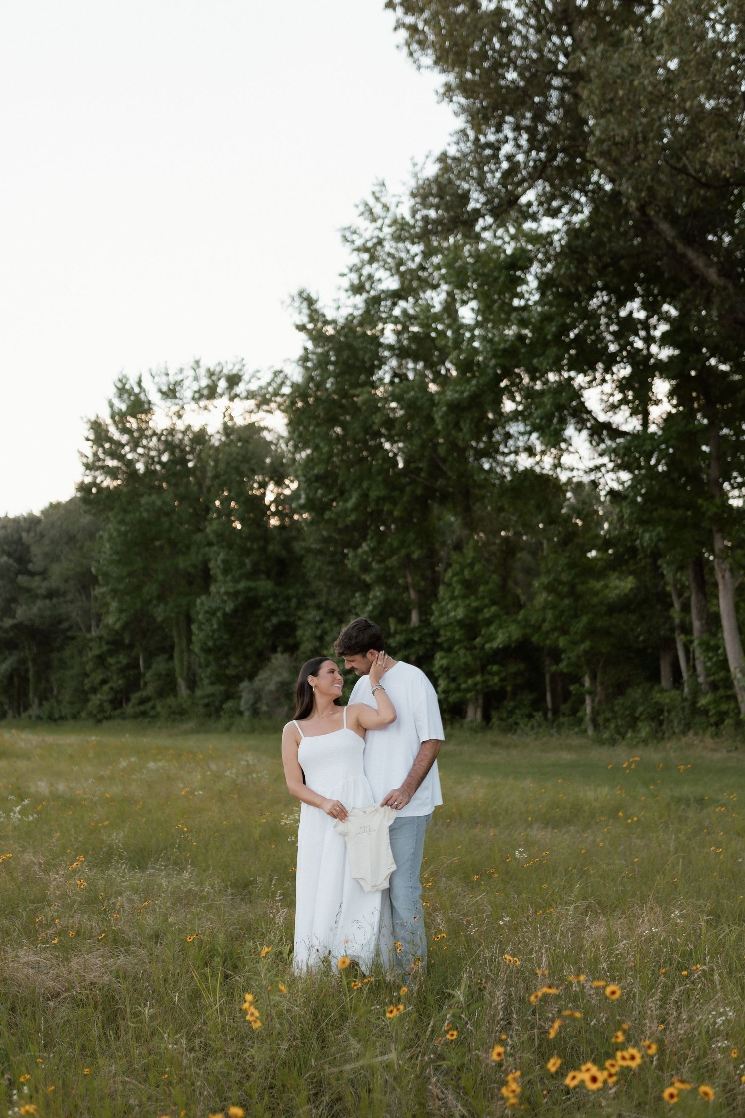 Couple standing in a wildflower field, woman in white dress with baby bump, man in white tee and jeans, surrounded by trees.