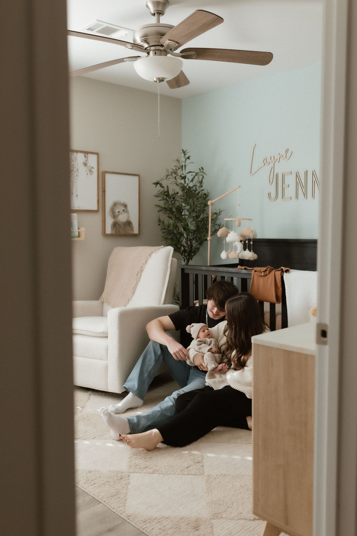 New parents sit on the nursery floor holding their newborn near a crib with personalized name wall decor.