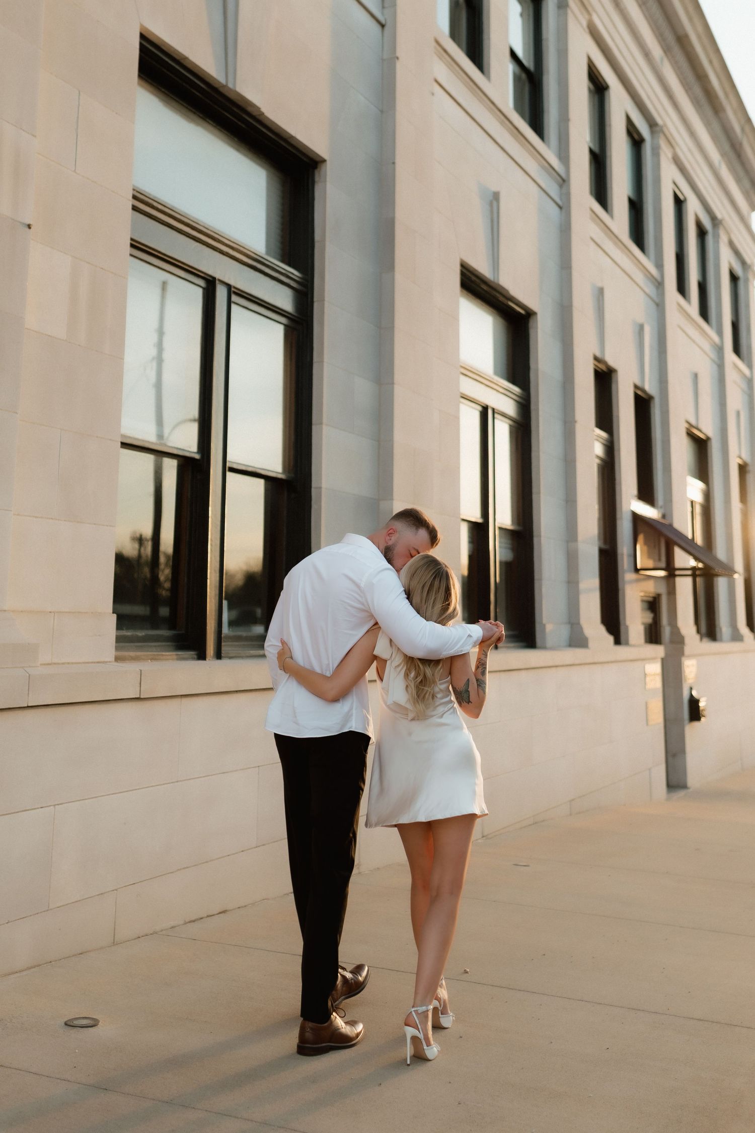 A couple walks arm in arm along a sunlit neoclassical building facade at golden hour.