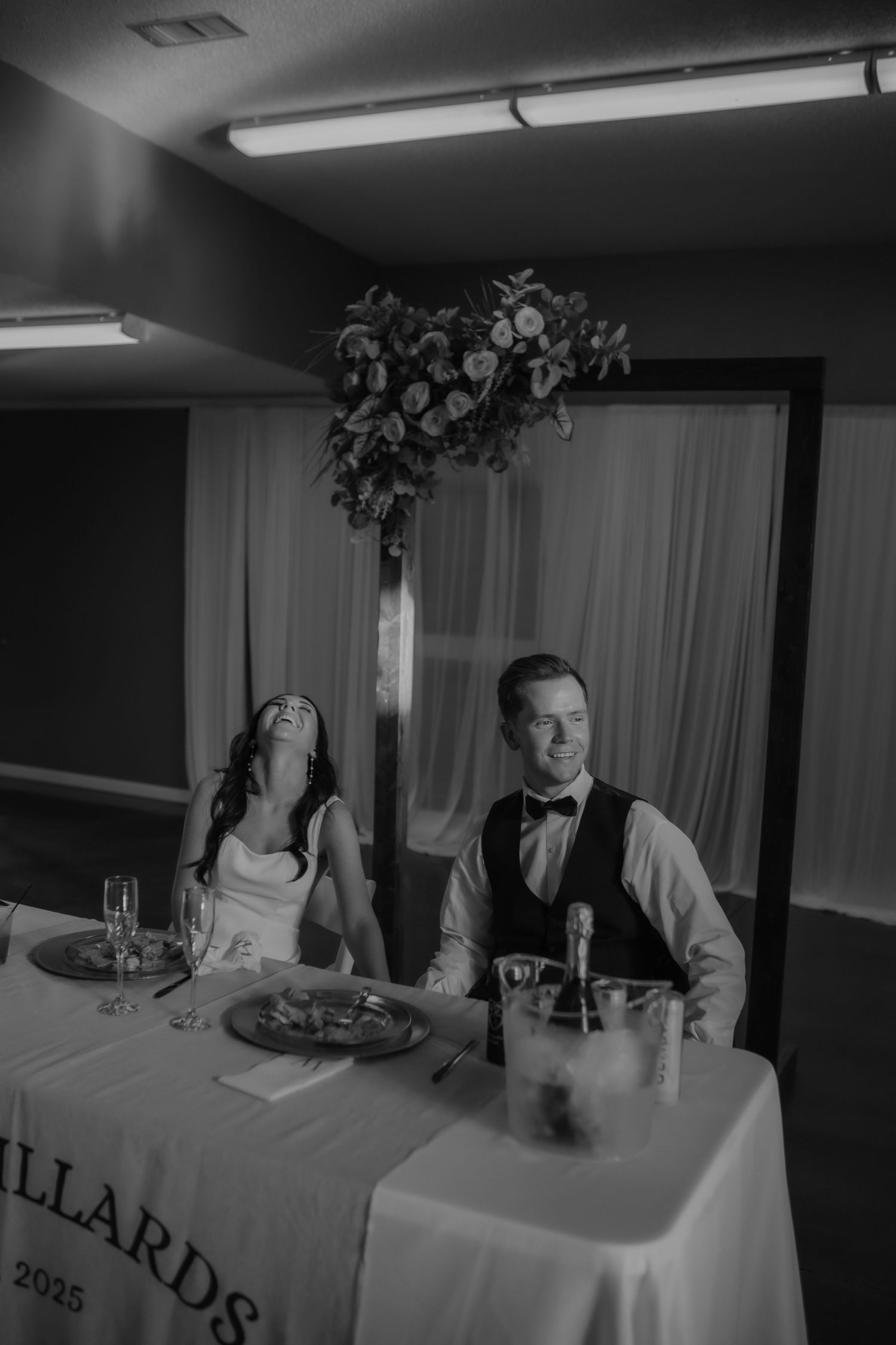 Bride and groom share a laugh at sweetheart table during wedding reception dinner.